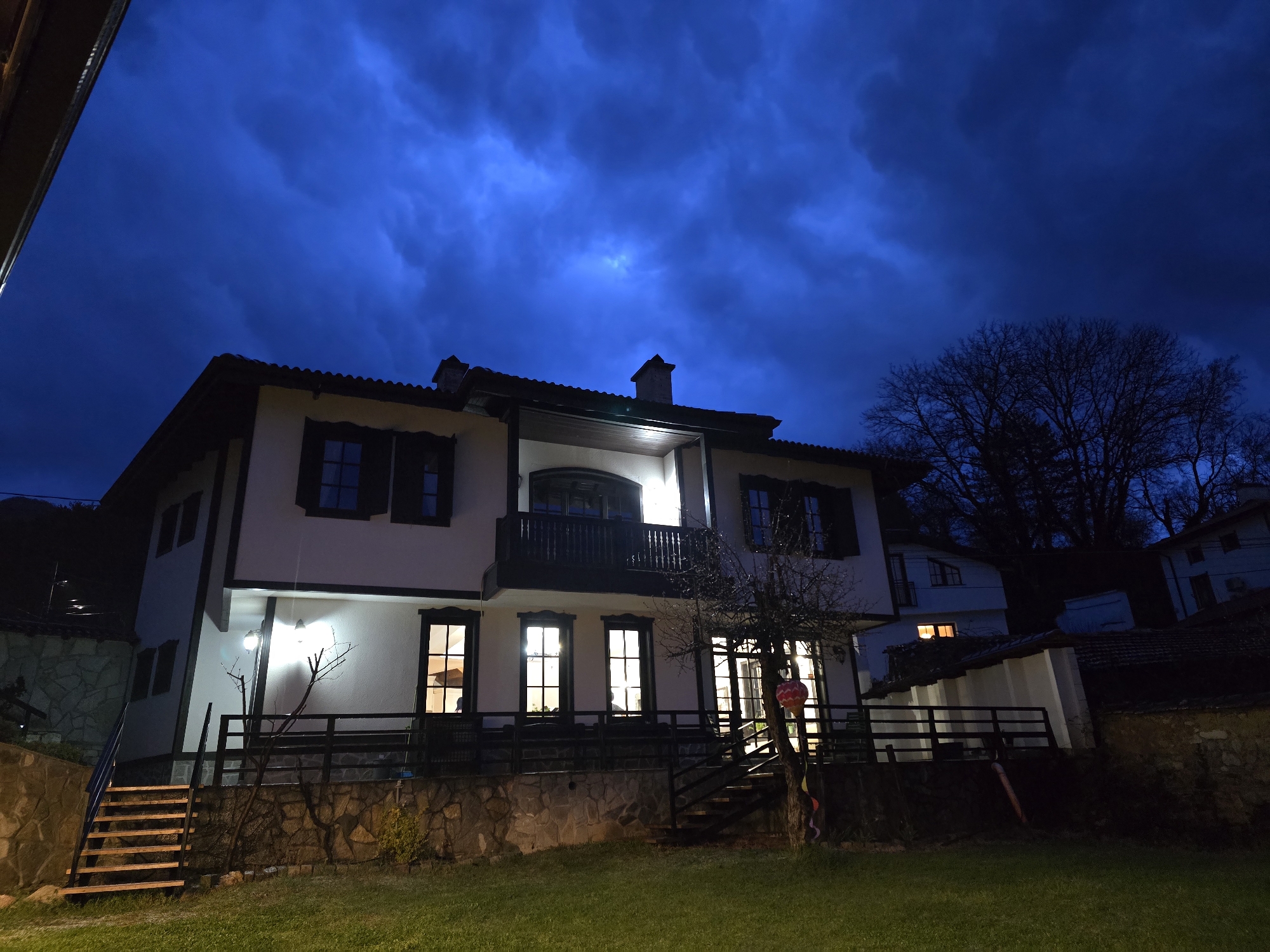 The image displays a two-story residential building with a white facade and dark brown architectural trim, illuminated at night under a dark blue, cloudy sky. The house features a tiled roof with visible chimneys, and its lower section is constructed with light brown and gray stone blocks. Multiple windows with dark frames are visible; several on the ground and second floors are lit from within, emitting a warm yellow light. Some upper-story windows are equipped with dark shutters. A prominent balcony with dark railings extends from the second floor, located above a brightly lit entrance area. Several exterior wall-mounted lights illuminate the facade. In the foreground, a well-maintained green lawn is visible. To the left, wooden steps with a black metal handrail ascend towards the house. To the right, a bare deciduous tree, decorated with a red-orange lantern and colorful ribbons, stands adjacent to a dark fence or railing system that encloses a ground-level patio area. In the background, to the right of the main house, a portion of another building with an illuminated window is visible, along with silhouetted trees and distant landforms. No individuals or animals are present. The location is Unknown, Unknown.