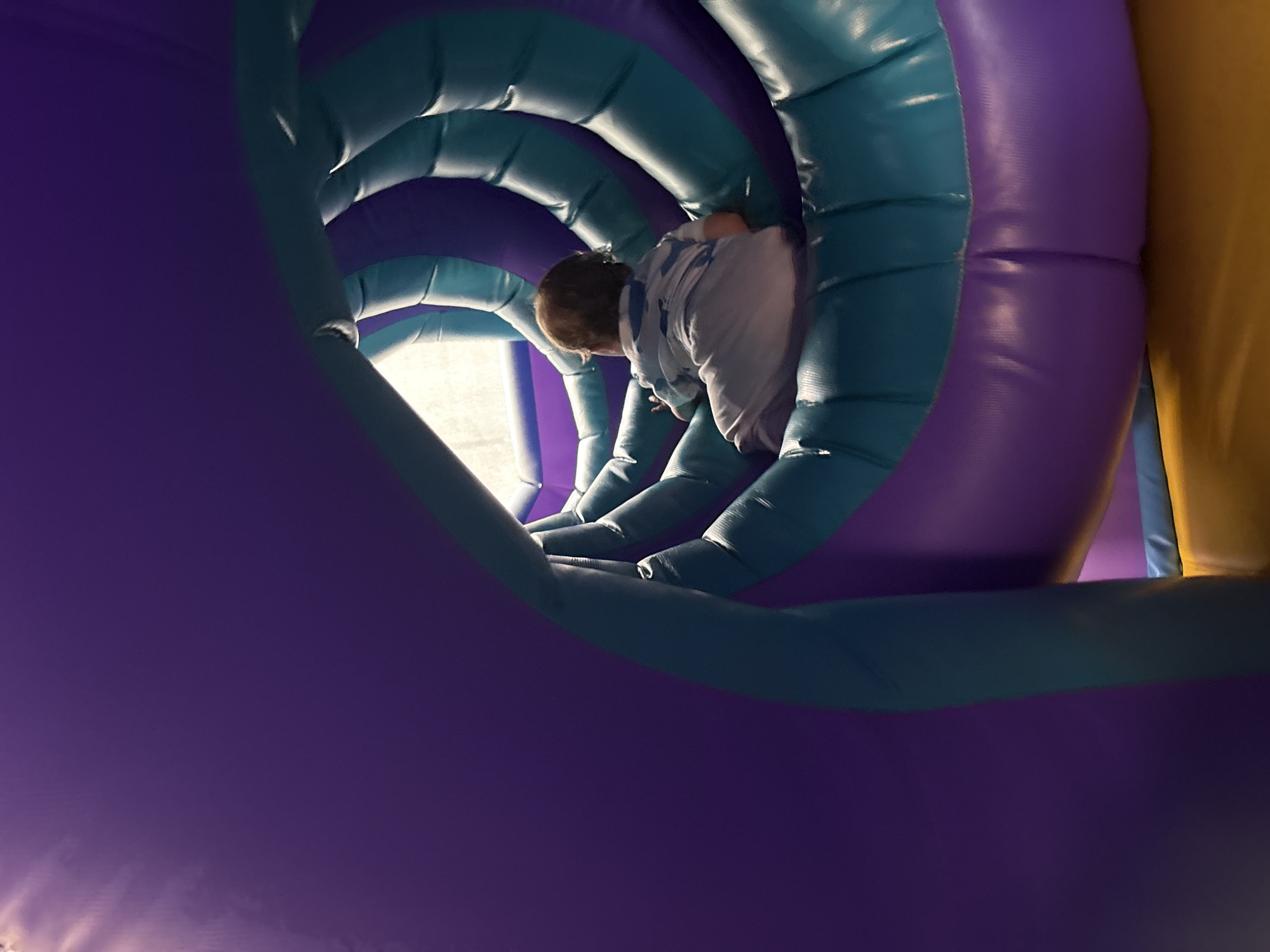 A child is crawling through a purple and teal inflatable tunnel. The tunnel is curved, creating a swirling effect. The child is wearing a white shirt. The background shows a bright light source at the tunnel's exit. The exact location within Izhevsk, Russia, cannot be determined from the image.