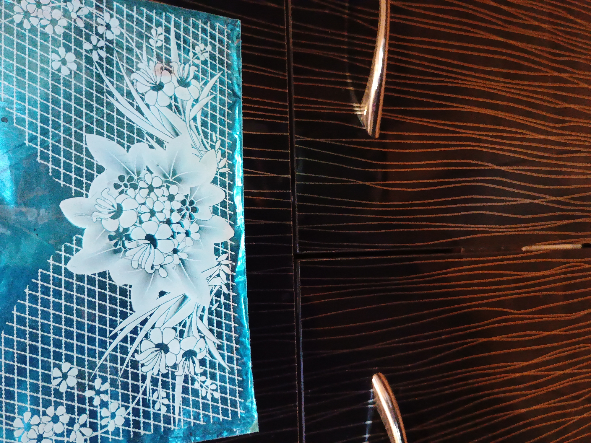 The image displays a close-up of a dark, possibly wooden, cabinet with two doors. Each door has a metallic, curved handle. The top portion of the image shows a decorative frosted glass panel with a floral design against a bright blue background. The glass panel is positioned above the cabinet doors. The cabinet itself has a textured surface with vertical, wavy lines in a lighter brown or orange hue against the dark background. The scene is captured indoors. There is no indication of a specific location within the city of Zaporizhzhia, Ukraine.