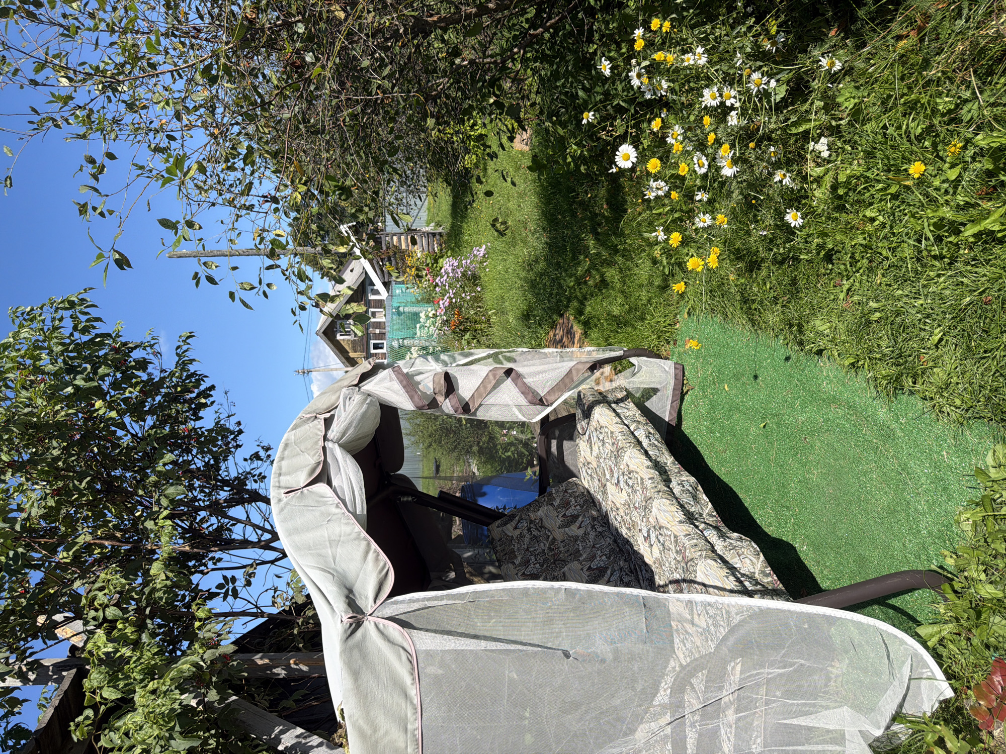 The image depicts a sunny outdoor scene in Yalguba, Russia, likely a backyard. In the foreground, a fabric-covered garden swing is set on a patch of artificial green turf. The swing is draped with a white mosquito net, and its cushions have a patterned upholstery. To the right of the swing, a cluster of white and yellow wildflowers grows in the grass.
In the background, several wooden houses with gabled roofs are visible, suggesting a rural or semi-rural residential area. A green fence separates the properties, and beyond that, trees and vegetation are abundant. The sky is a clear, bright blue. The overall impression is one of a quiet, summery day in a Russian dacha environment. No people are present in the image.