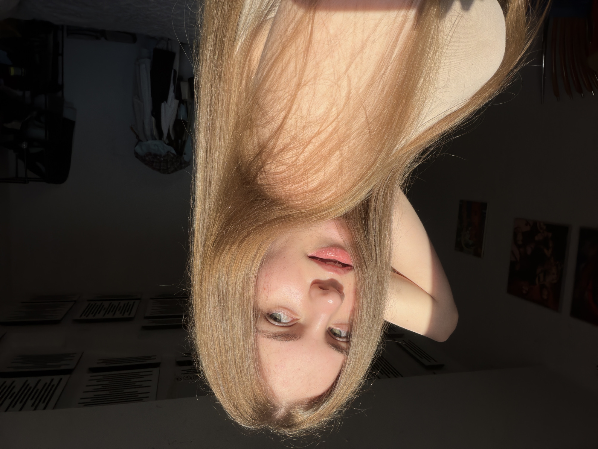 The media file is a portrait of a young person with blonde hair, taken from a slightly elevated angle, as if the camera is held above their head. The subject's head is tilted downwards, so their face is positioned towards the bottom of the frame. They have light-colored eyes, rosy cheeks, and their lips are slightly parted, appearing to have a gloss or balm on them. Their hair is long and flows around their face and shoulders, catching the light. The subject is wearing a beige top.
The background suggests an indoor setting, possibly a bedroom, with furniture and decor visible. To the left, there appears to be a closet or wardrobe with clothes hanging inside. Further back, on the wall, are several framed items, possibly artwork or posters, arranged in a grid-like pattern. The lighting indicates that it is daytime, with strong sunlight casting shadows and creating highlights on the subject and the environment. The presence of a closet and personal decor suggests a residential setting. The time of day appears to be afternoon or late morning, given the angle and intensity of the sunlight. No visible text is present in the media file. The overall mood is relaxed and intimate, likely a selfie. The location context provided is Dnipro, Ukraine.