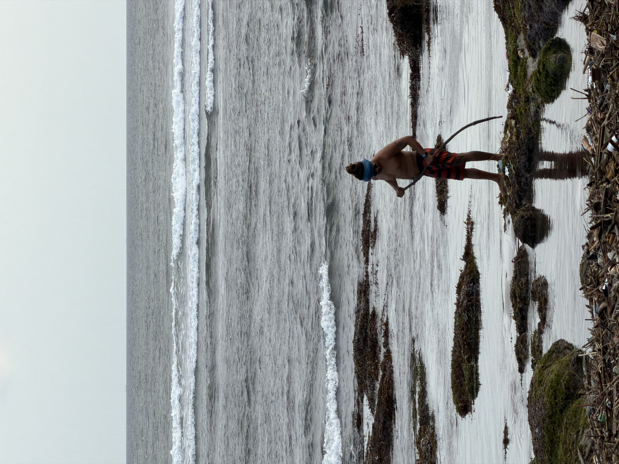 A shirtless man wearing red and black striped shorts and a blue headband stands on a rocky shore covered in seaweed and debris. He is holding a long, curved stick, possibly a tool for gathering or moving the seaweed. The ocean is calm with gentle waves rolling in. In the background, the horizon line separates the sea from an overcast sky. The foreground shows more rocks, seaweed, and a collection of washed-up driftwood and trash, indicating a natural, somewhat untended shoreline. The scene suggests a man engaged in some form of activity related to the coastal environment. This location is consistent with the beaches of North Kuta, Indonesia, which are known for their natural beauty and sometimes rugged coastlines.