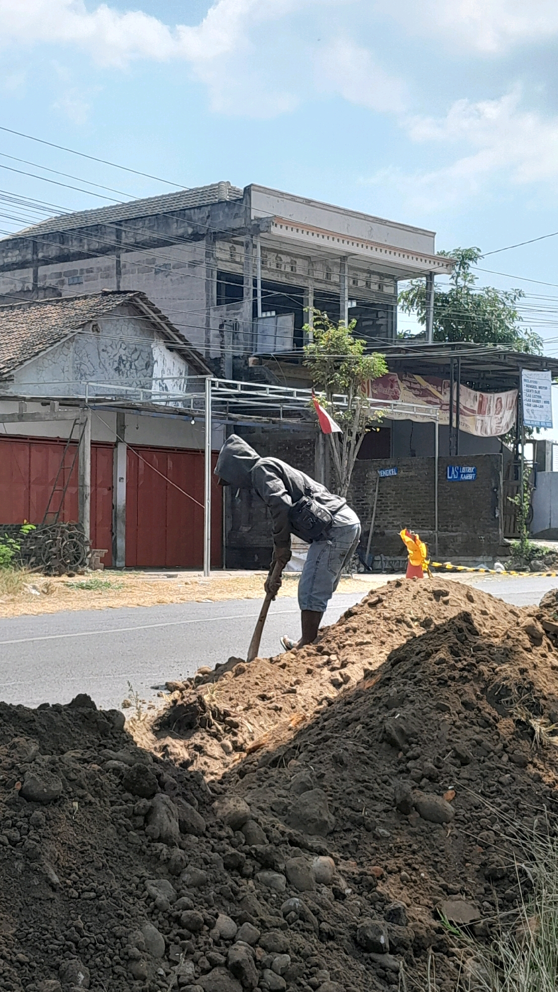 In Tanjung, Indonesia, a construction worker, dressed in a grey hooded jacket, blue jeans, and dark boots, is actively digging a ditch on the side of a paved road. They are using a shovel with a wooden handle to move a pile of dark soil and rocks. The immediate foreground is dominated by this large mound of excavated earth. In the background, a two-story building with a grey tiled roof and unfinished construction elements is visible. A red metal door and a ladder are present on the left side of the building. Further down the street, a building with a corrugated metal roof features signage and a red and white Indonesian flag. A bright yellow object, possibly a traffic cone or safety marker, stands near the road's edge. The sky above is clear with scattered white clouds, and overhead power lines are visible.