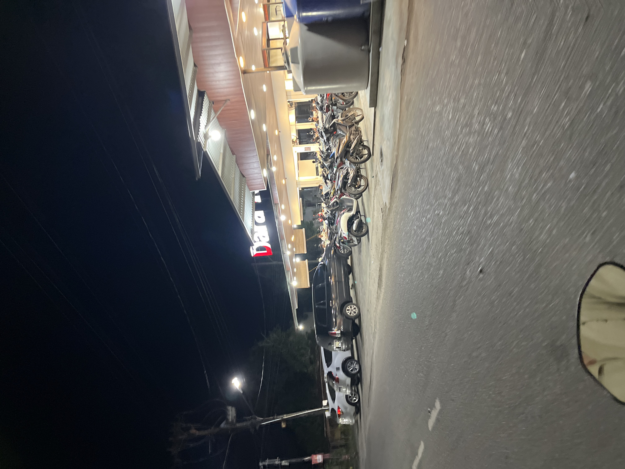 The image shows a nighttime scene in front of a building with a sign that reads "Dara". The building appears to be a commercial establishment, possibly a store or restaurant, with a covered outdoor seating area or awning. A street is in the foreground, with a white line painted on it. To the right of the street, there is a parking area filled with motorcycles. Behind the motorcycles, several cars are parked. The building itself is lit up with numerous lights, and the overall atmosphere is that of a busy commercial area. The location is identified as being in the city of Langsa, Indonesia.