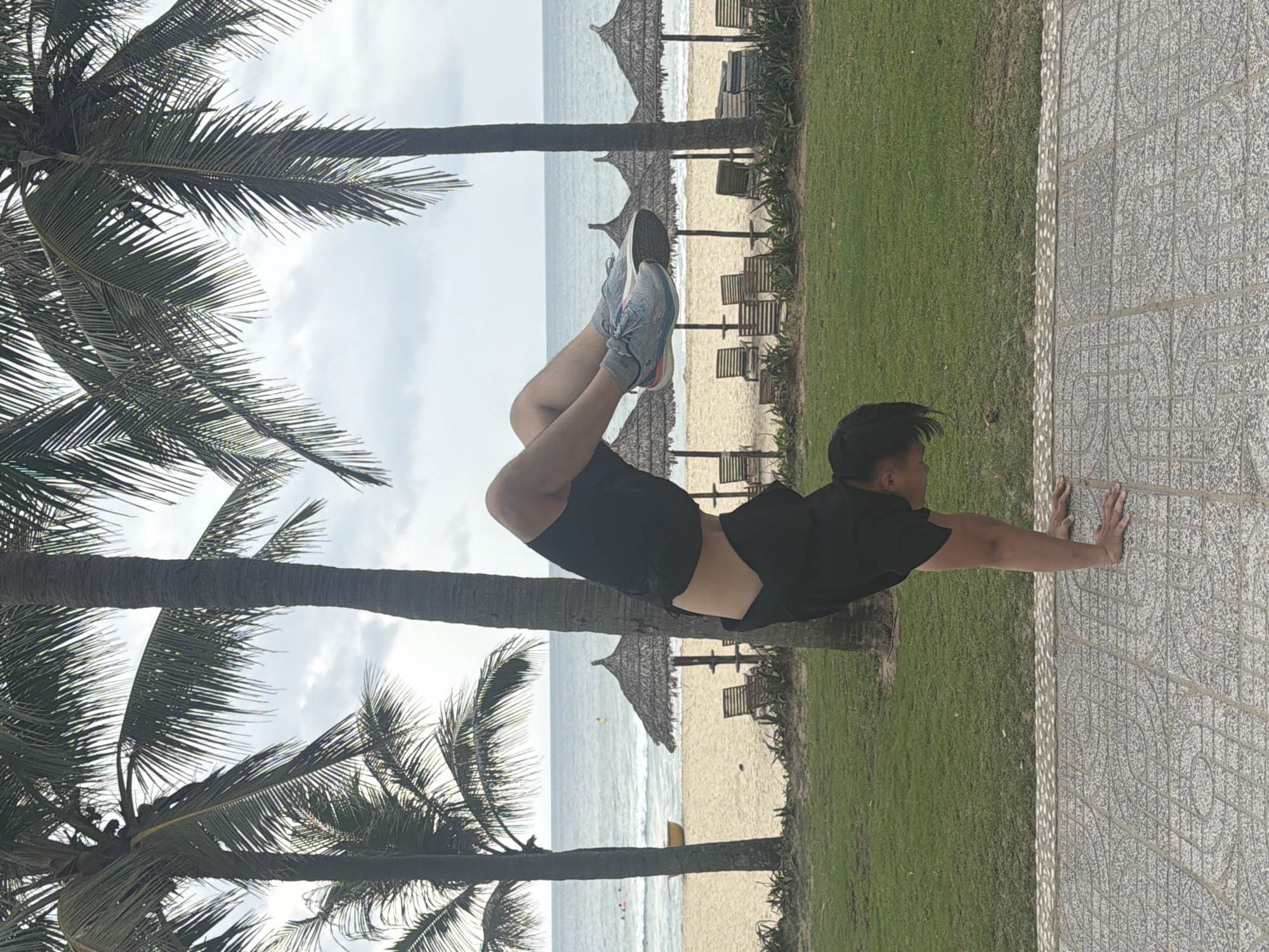 An individual, likely male, is performing an impressive horizontal arm balance pose. Their body is suspended in the air, parallel to the ground, supported by their hands placed firmly on a patterned tiled path. They are wearing a black short-sleeved t-shirt, black shorts, and light-colored sneakers with pink and blue accents on the soles. Their legs are bent at the knees and lifted, contributing to the balance.
The scene is set outdoors, likely at a beach resort in Đà Nẵng, Vietnam, under an overcast sky. In the background, beyond the tiled path and a strip of green grass, lies a sandy beach. Several large palm trees with dense fronds frame the left side of the image and are visible throughout the background. Thatched beach umbrellas and wooden lounge chairs are scattered across the beach. The calm ocean stretches out to the horizon behind the beach. The tiled path features a repetitive geometric pattern in shades of grey. No visible text is present in the image.