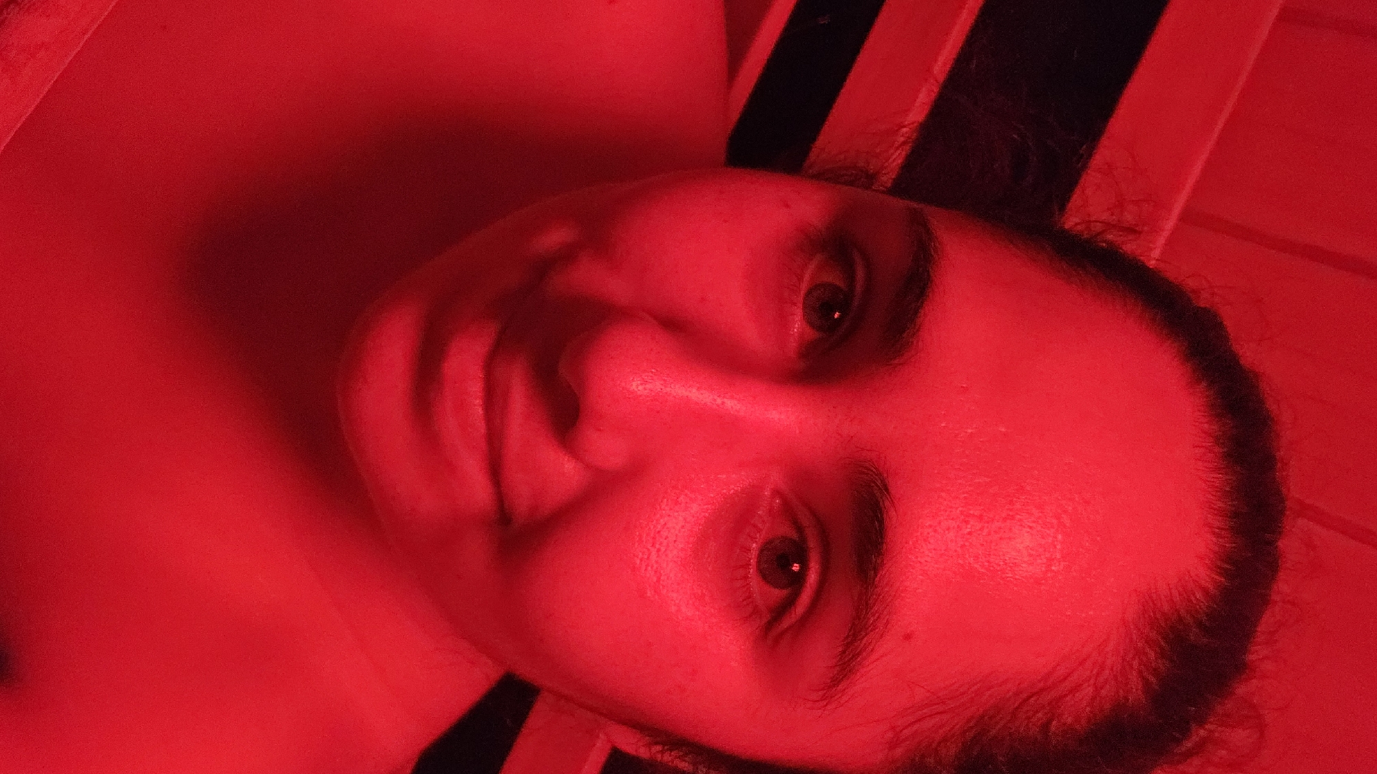 An individual, likely a young adult woman, is depicted in a close-up, head-and-shoulders portrait, lying down and looking directly at the viewer. The entire scene is saturated in a strong, monochromatic red light, casting a vivid crimson hue over everything. The person's face is central, with open eyes whose dark pupils and irises contrast against the reddish sclera. Her facial features, including her nose, lips, and eyebrows, are clearly visible, rendered in shades of red and shadow. Dark hair frames her face, discernible against the red background. She appears to be resting on a surface that features prominent black stripes, visible behind her head and extending to the upper right of the frame, possibly part of a bed, chair, or patterned wall. The overwhelming red illumination creates a dramatic, intense, and somewhat surreal atmosphere. Her expression is neutral and calm. There are no other objects visible, nor any discernible text. The setting appears to be an indoor environment, possibly a red light therapy room or a space designed with specific mood lighting, located in Puebla, Mexico.