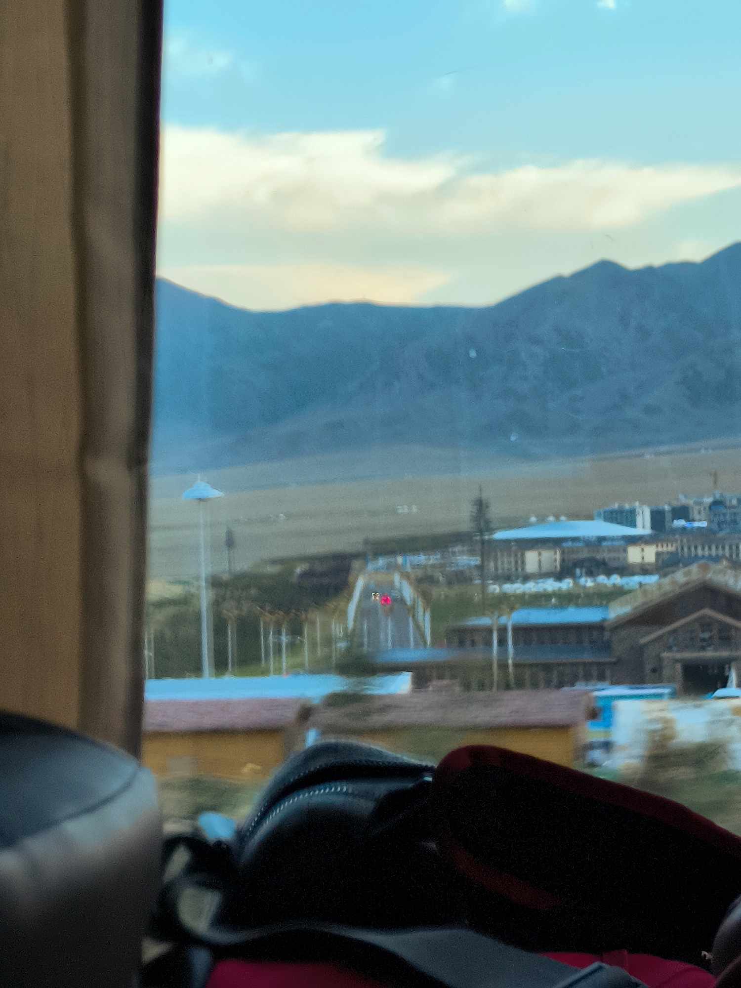 The image, captured from inside a moving vehicle, shows a blurred view through a window overlooking a vast, mountainous landscape and a town. In the foreground, partially obscuring the view, are dark, soft objects, likely bags or luggage, with a red edge visible on one. Beyond the immediate foreground, the scene opens up to a town with various buildings, some with blue roofs, others with tan or darker brown facades. A tall, slender white structure, possibly a lamppost or a monument, stands prominently in the mid-ground. In the distance, rolling hills and majestic mountains form the backdrop under a pale blue sky with scattered clouds. A small flag, red with a yellow symbol, is visible on a structure within the town, indicating a possible location in China. The overall impression is one of travel and a panoramic view of a rural or semi-rural settlement against a dramatic natural landscape.
