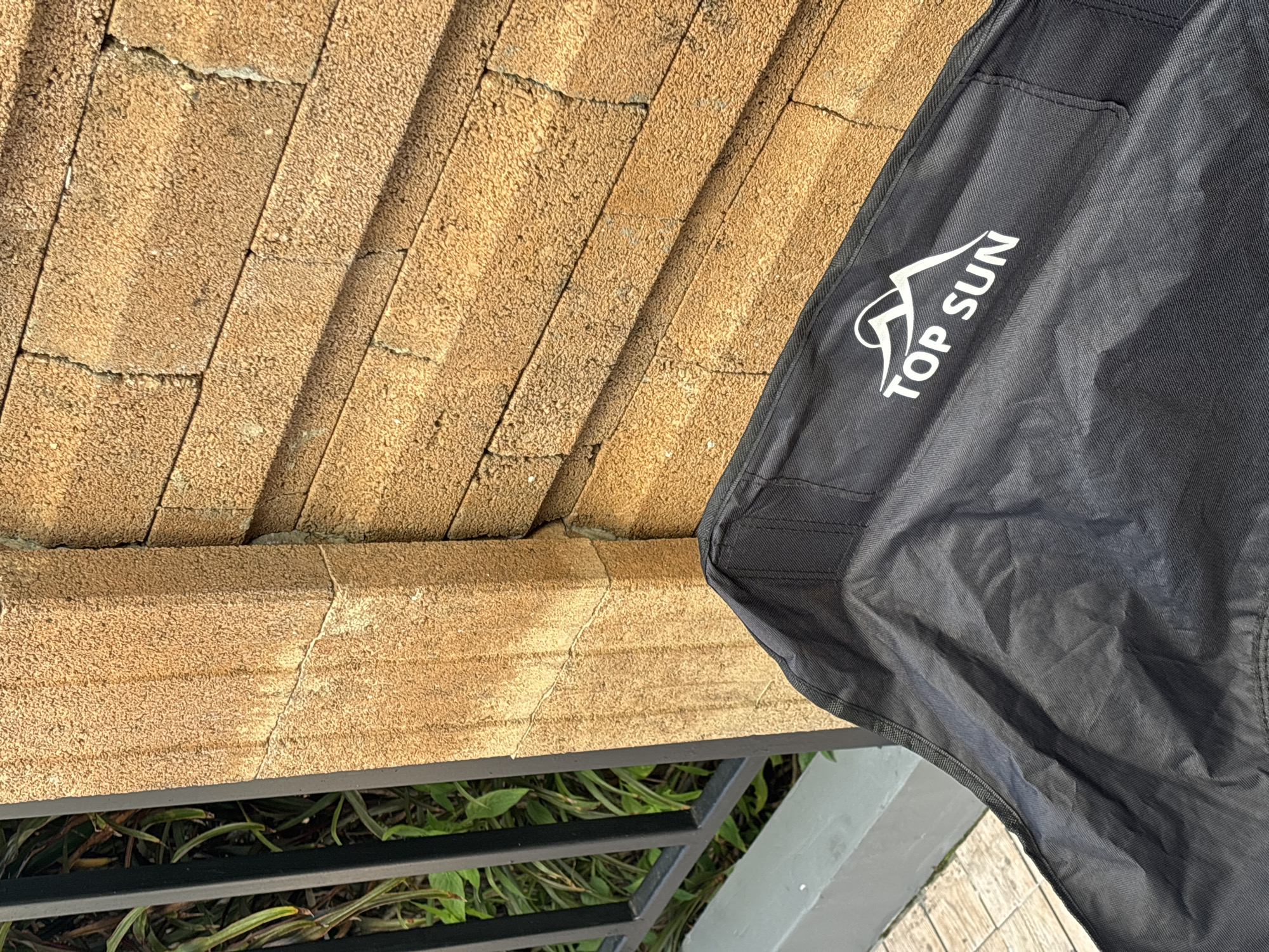 The image is an outdoor, medium-shot, eye-level photograph taken at eye level. It features a black fabric object, possibly a bag or tarp, with the white logo "TOP SUN" and a mountain silhouette printed on it. This object is in the foreground, partially obscuring a light brown brick wall. The wall appears to be constructed of rectangular bricks with a rough, textured surface, and some grout lines are visible. To the left of the brick wall, a black metal fence with vertical bars is visible, with green foliage behind it. In the lower left corner, a light grey concrete ledge is present. The scene suggests an outdoor setting, likely in a residential or commercial area in Ko Kaeo, Thailand, given the context provided. The lighting indicates it is daytime, with sunlight casting shadows.