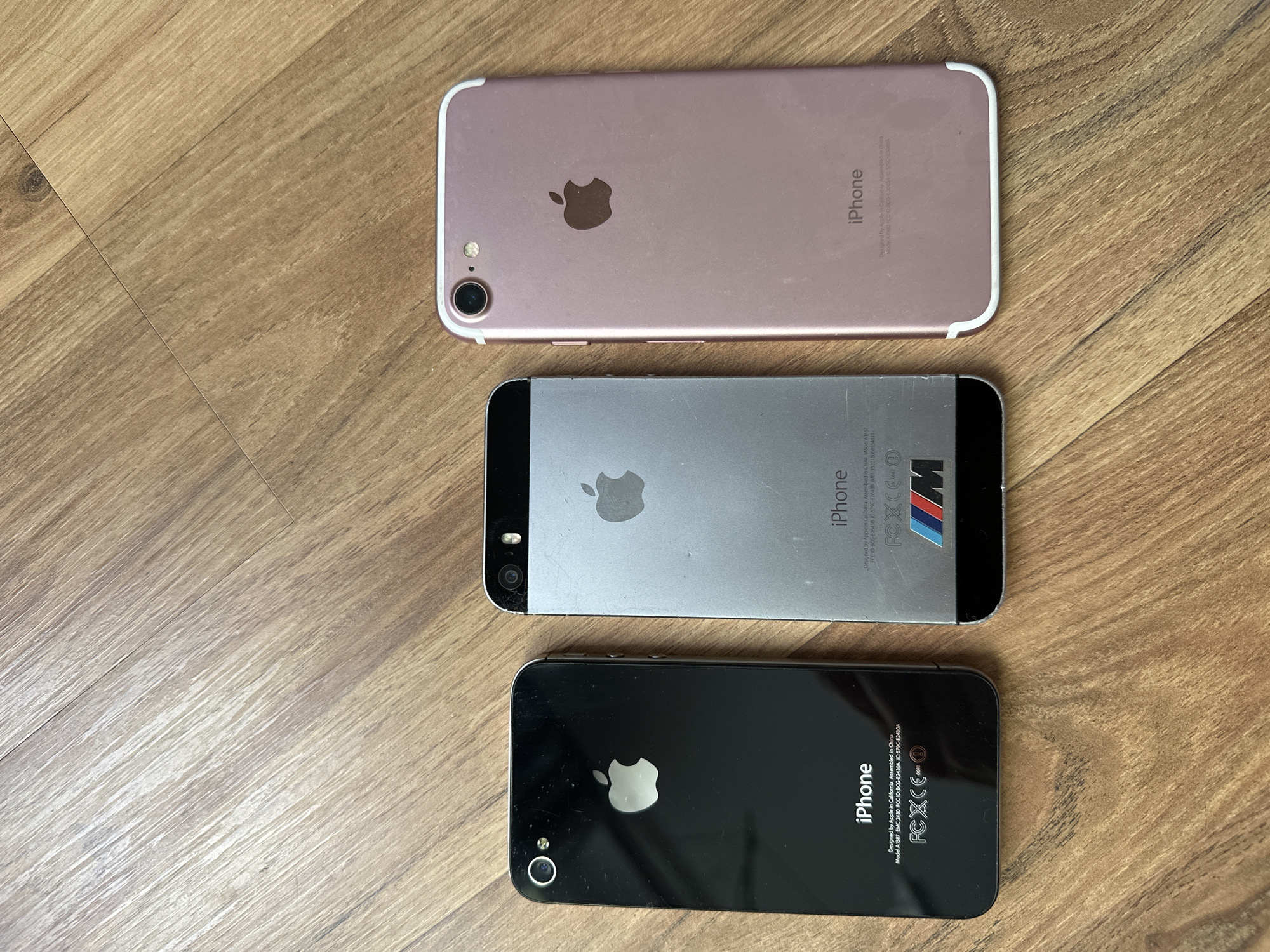 Three Apple iPhones are positioned horizontally on a light brown wooden floor, displaying their rear casings. From left to right: a black iPhone 4S (Model A1387) shows a glass back with a circular camera lens and flash; a silver/space gray iPhone 5S (Model A1457) features a two-tone metal and glass back with a circular camera lens and flash, and a multi-colored BMW M-series logo sticker affixed to its lower half; and a rose gold iPhone 7 (Model A1660) displays a metal unibody back with a larger circular camera lens and oval-shaped flash, and antenna lines integrated into the top and bottom edges. All devices exhibit minor surface dust and wear consistent with use. The wooden floor planks run diagonally across the frame from bottom-left to top-right. The scene is located in Unknown, Unknown.