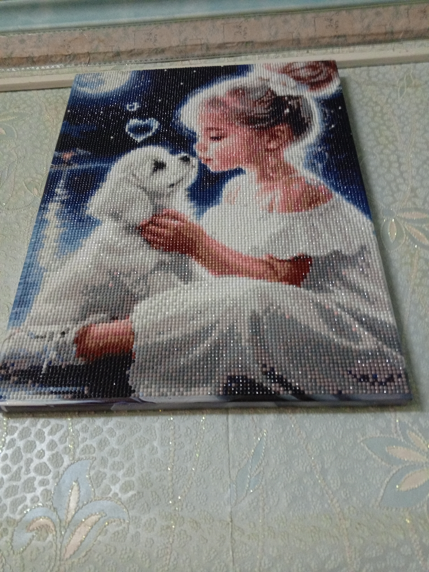 A close-up, slightly angled, indoor shot shows a completed diamond painting depicting a girl embracing a white dog. A heart shape is visible above the dog's head. The background of the painting features a dark blue hue interspersed with white specks. The painting rests on a light-colored surface with a subtle floral pattern. The background is slightly out of focus, displaying a wall or similar surface with a pale blue and beige pattern. The location is Puchezh, Russia.