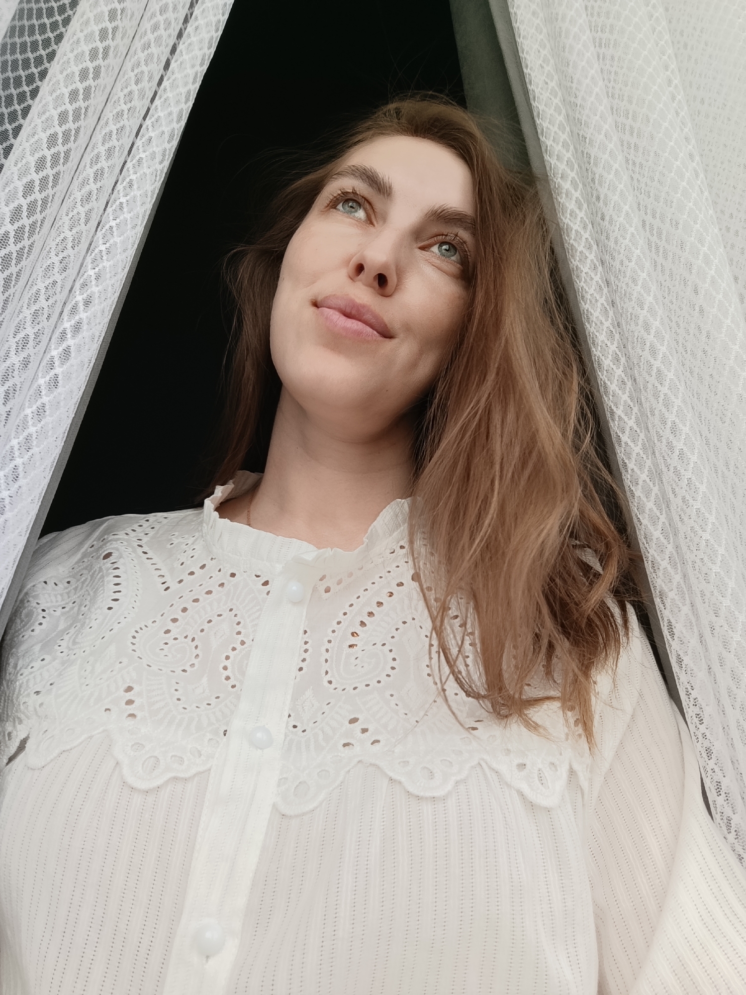 A woman with light brown hair and fair skin is looking upwards, framed by white lace curtains. She is wearing a white, long-sleeved blouse with a decorative lace collar and button-down front. The background appears dark, possibly an open window or doorway. The overall setting suggests an interior space. There is no evidence to suggest a specific location, such as the city of Chaikovsky, Russia.