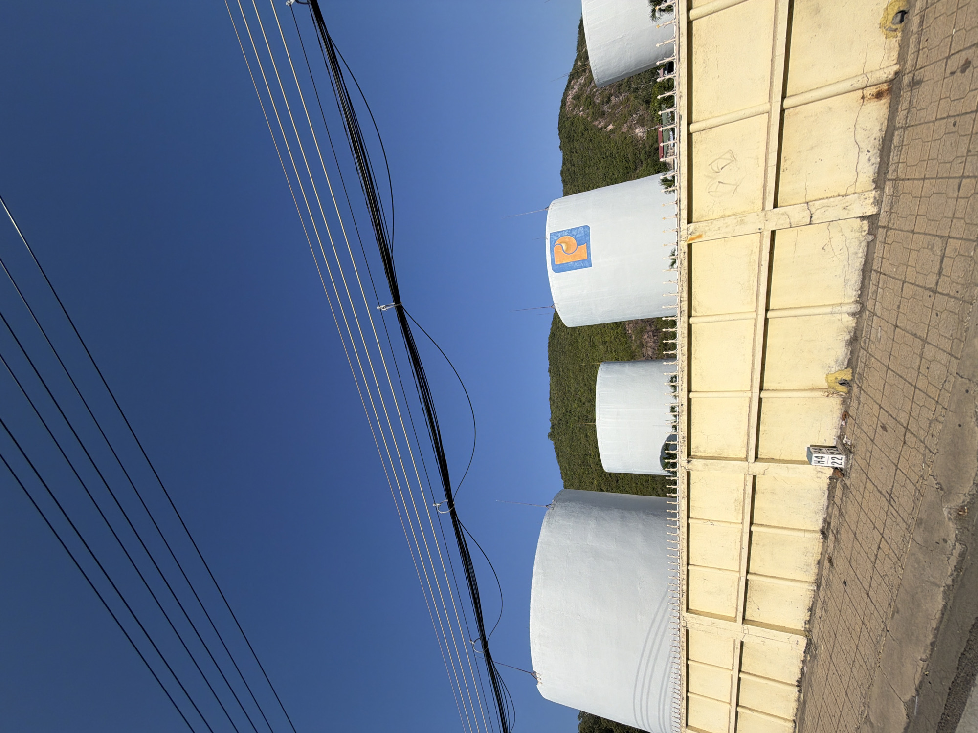 The image depicts an industrial scene in Nha Trang, Vietnam, under a clear blue sky. In the foreground, a weathered yellow concrete fence runs horizontally across the frame, topped with decorative white spikes. The fence is built with rectangular panels and shows signs of age, with cracks and discolored patches. Beyond the fence, four large, white cylindrical storage tanks dominate the middle ground. The tank on the far left is the largest, followed by two progressively smaller tanks to its right. The fourth tank, partially visible on the far right, is also white and cylindrical. One of the middle tanks features a logo composed of a blue square with an orange “P” inside. The tanks are situated on a slight incline, with a green, tree-covered hillside rising behind them. Overhead, multiple black and thin white electrical wires are stretched across the sky, suspended by poles not fully visible in the frame. The ground in front of the fence is paved with light-colored square tiles arranged in a grid pattern, with a concrete curb bordering the lower edge of the image. A small, white marker with black markings, possibly a survey or identification marker, is visible near the center of the fence. The overall impression is one of an operational industrial area against a natural backdrop.