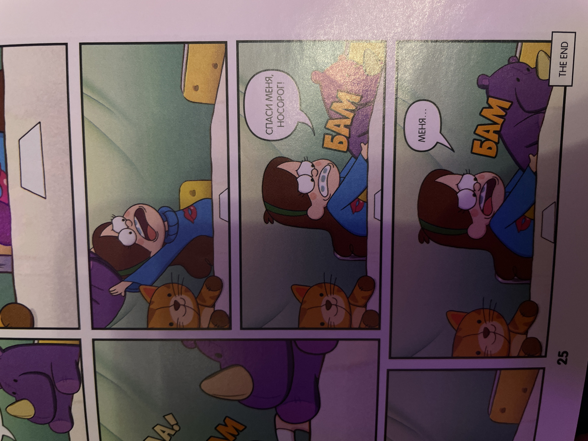 The image is a comic book panel, specifically page 25, which depicts a scene from the animated series Gravity Falls. The panel is divided into four smaller rectangular frames.
In the top left frame, a stuffed purple rhinoceros toy is visible. The top right frame shows a young girl with brown hair, wearing a blue sweater with a red and yellow emblem, crying and holding a stuffed purple rhinoceros. A stuffed orange cat is to her left, and a yellow rectangular object, possibly a cheese slice, is to her right.
The bottom left frame also shows the stuffed purple rhinoceros and a stuffed orange cat. The bottom right frame shows the same girl, still crying, holding the stuffed purple rhinoceros. The speech bubble above her head reads "МЕНЯ..." (MENYA...), which translates to "ME...". Below this, a large sound effect reads "БАМ" (BAM) in yellow letters. A stuffed purple bear is lying on its side next to the rhinoceros. The frame also contains a small box labeled "THE END".
The background in the comic panels is a gradient of green and light blue, suggesting an indoor environment. There are no discernible elements of the city of Sinyavino, Russia, present in the image.