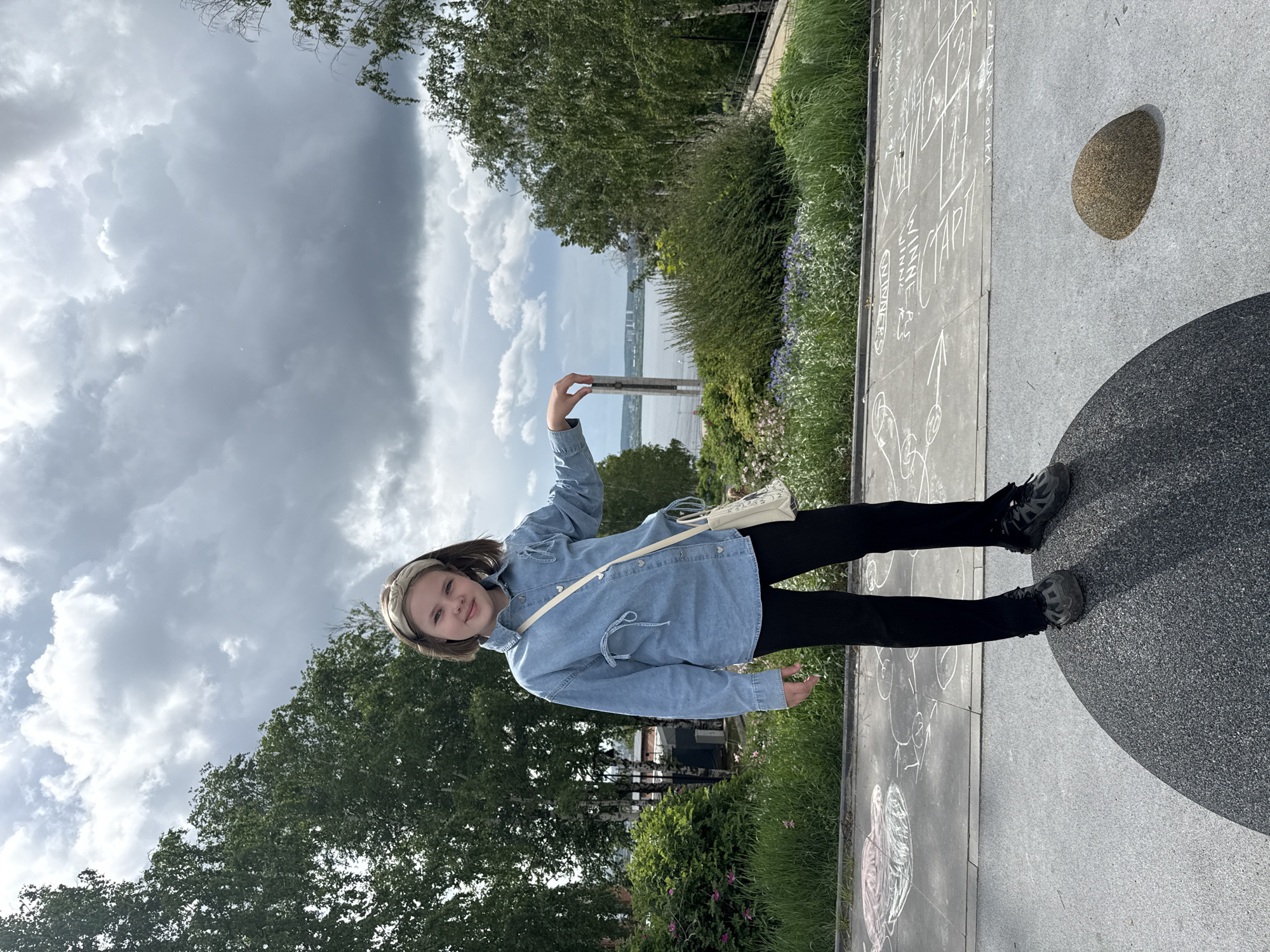 A young girl with light brown hair and a light headband stands smiling, posing for a photograph in an outdoor setting in Izhevsk, Russia. She wears a light blue denim jacket over black leggings and dark sneakers, with a light-colored shoulder bag draped across her body. Her right arm is slightly raised.
The scene is a paved area, possibly a promenade or park, under an overcast sky with large grey and white clouds. In the background, lush green trees frame a wide body of water, likely a river or lake, with distant structures visible on the opposite shore. The ground around the girl features light grey pavement, partially covered with numerous chalk drawings, including outlines of a hopscotch game, hearts, and arrows. Visible text in chalk includes words like "WINNIE," "MINE," "TAPPI," "OIKA," and the number "55," among others. To her right, a speckled, round stone object rests on the pavement, adjacent to a large, dark circular paved area. The overall impression is a daytime scene with soft, diffused light due to the cloudy weather.