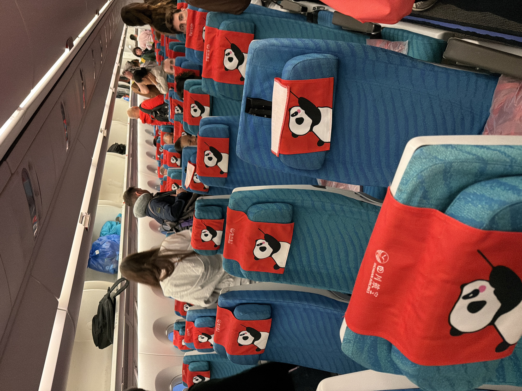 This media file, oriented vertically, captures the interior of an airplane cabin, likely an economy or standard class section, during what appears to be a flight or boarding process. Rows of blue, textured airline seats extend into the background, with the aisle visible to the left. The cabin is well-lit, suggesting either artificial lighting or daytime conditions.
Numerous passengers occupy the seats, mostly facing away from the camera, though some faces are partially visible. Passengers are casually dressed, with one person in the mid-ground wearing a neck pillow.
A prominent detail across the seats are red fabric headrest covers, each featuring a distinct cartoon panda design. Some pandas appear to be hanging, while others are standing or walking. The headrest covers also display white text: "四川航空" (Sichuan Airlines) and "SICHUAN AIRLINES", indicating the airline operating the flight.
Overhead bins line the cabin ceiling to the left, with some partially open, revealing stored luggage such as a black backpack and blue plastic bags. The overall scene depicts a typical commercial flight environment, with passengers settled in their seats. The strong branding suggests the flight is related to Sichuan, China, known for its pandas. No specific emotions are discernible among the passengers, who generally appear calm.
