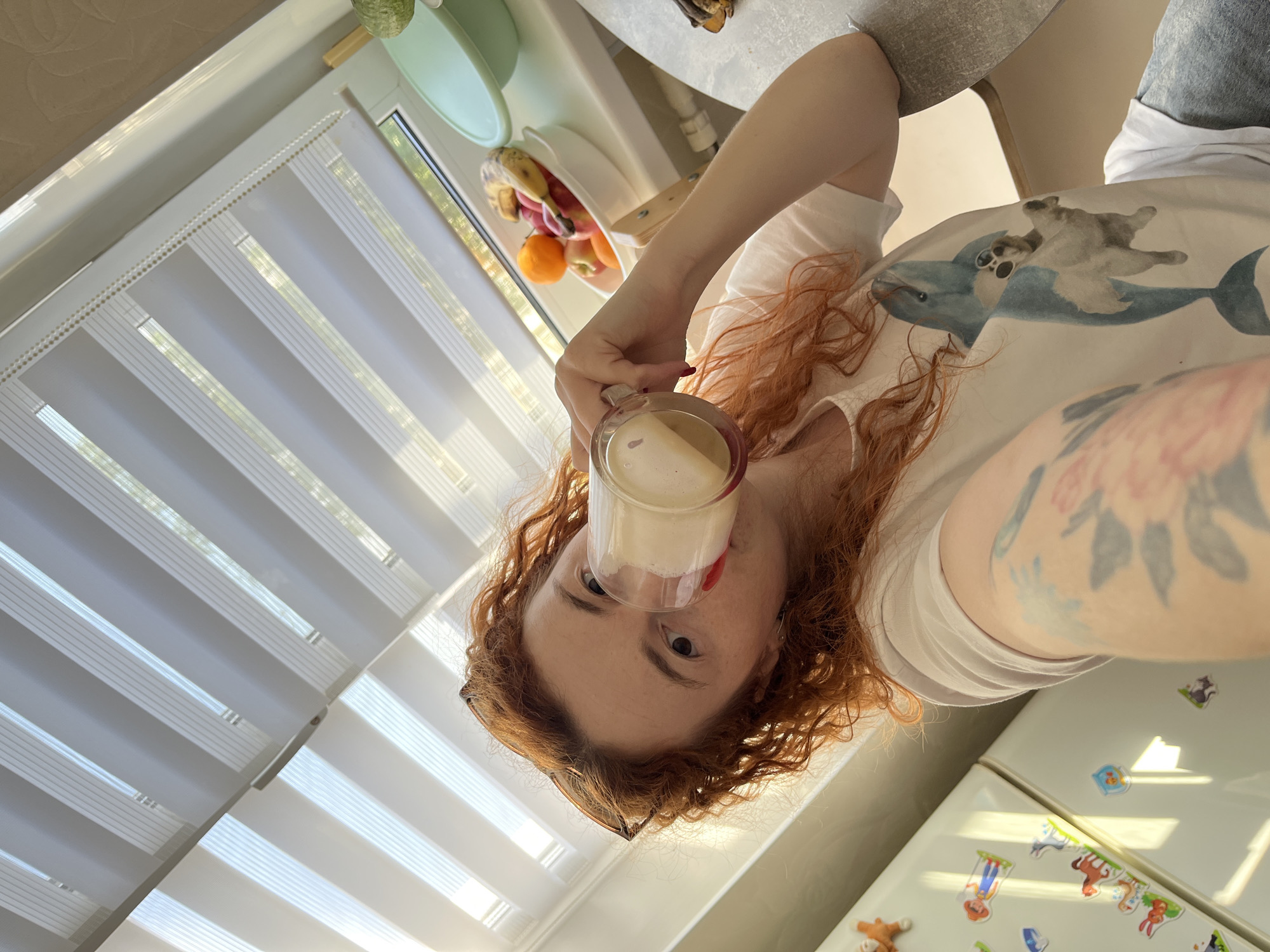 A woman with long, curly red hair is drinking from a glass mug filled with a light-colored beverage. She is wearing a white t-shirt with an image of a dolphin and a dog. The woman is indoors, likely in a kitchen, as evidenced by the presence of a refrigerator with various cartoon stickers on it. There is a window with white blinds in the background, and a fruit bowl is visible on the counter. The city is Chaikovsky, Russia.