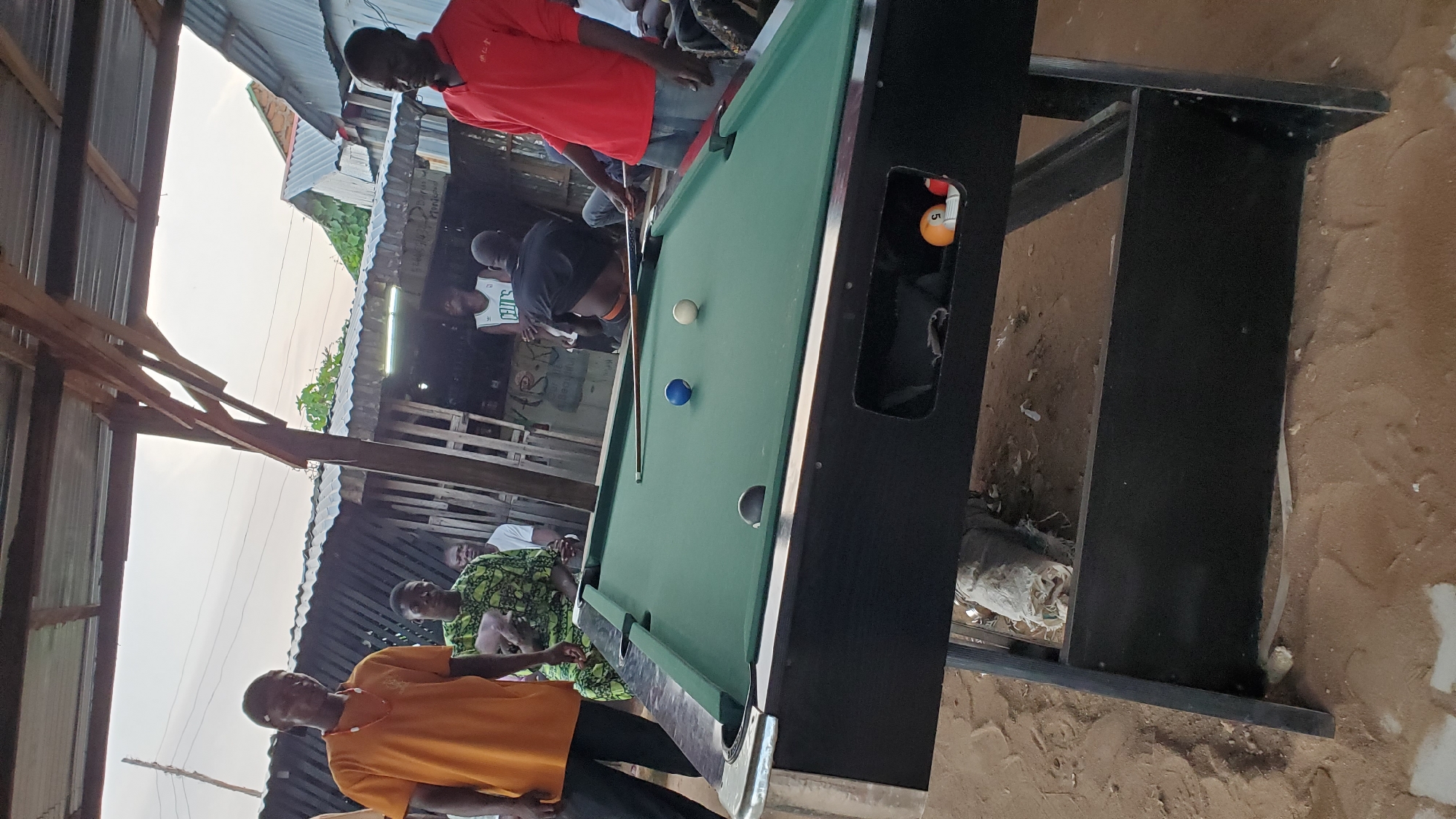 In the city of Jalingo, Nigeria, a group of individuals are gathered outdoors under a makeshift shelter. A green pool table dominates the foreground, with a cue stick resting across it. Three billiard balls are visible on the table: a solid blue ball, a solid white ball, and a solid black ball. The table is supported by a black, wooden frame, and beneath it, a compartment contains two more billiard balls: a striped ball with the number 5 and a red ball.
Several men are present. One man in an orange t-shirt and black pants stands to the left. Another man in a green and black patterned garment is seated. To the right, a man in a red long-sleeved shirt and blue jeans is standing, facing the pool table. In the background, a structure made of corrugated metal and wooden planks is visible, with some vegetation and other buildings. The ground appears to be sandy and uneven, with debris scattered around. The lighting suggests it is either early morning or late afternoon.