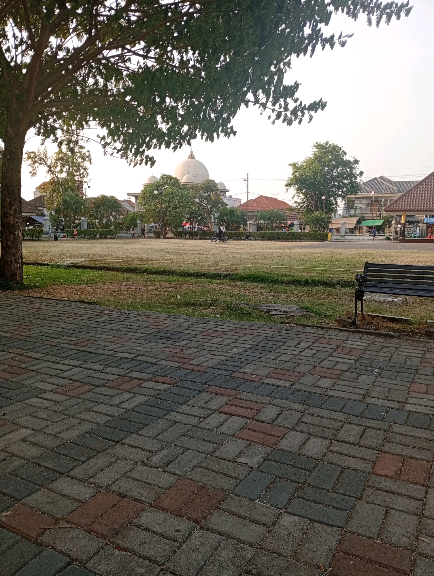 The image depicts a view of a park in Bangil, Indonesia, likely during the late afternoon or early morning given the soft light. In the foreground, a paved walkway made of rectangular bricks in various shades of grey and red herringbone pattern leads the eye towards the park. To the right, a dark metal park bench is visible.
The park itself is a large, open grassy area with some dry patches. A low, green hedge borders the grass. In the background, a white domed building, possibly a mosque or a public building, stands prominently. Several trees with green foliage are scattered throughout the park and beyond the hedge. Behind the trees and the domed building, there are residential buildings with tiled roofs.
Several people are present in the park. Two individuals are seen walking in the distance on the grass. To the left of the frame, near the trees, a person in an orange shirt is walking. On the far right, a person on a bicycle is moving away from the camera. The sky is overcast and bright, indicating a diffused light source.