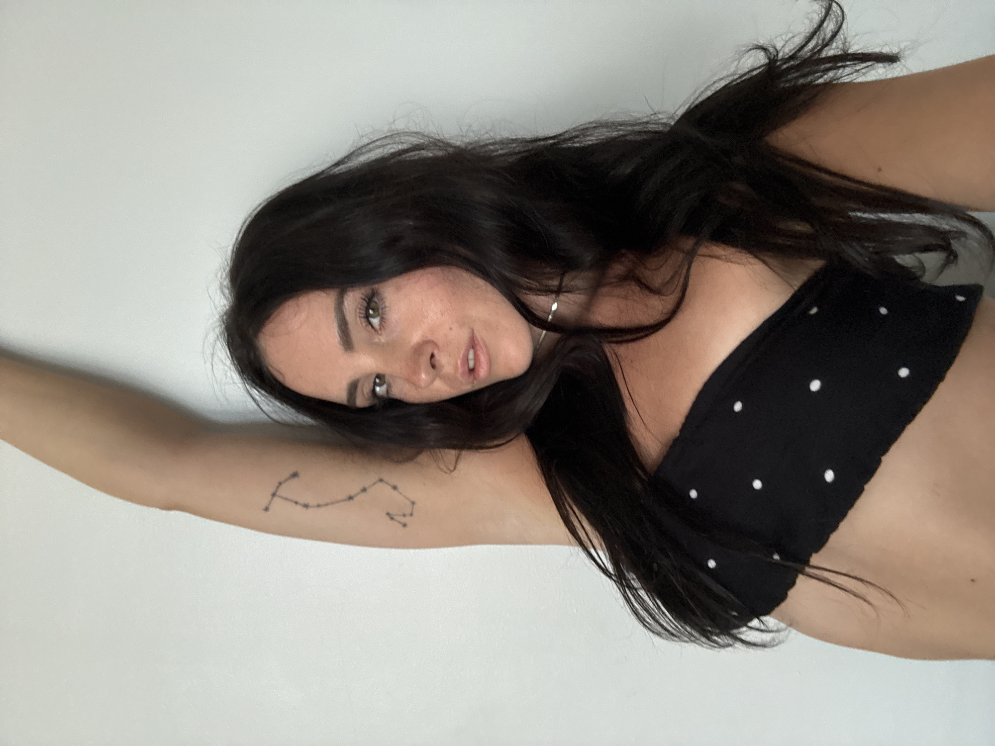 A woman with dark hair and a black polka dot bandeau top is shown from the waist up, with her left arm raised and bent at the elbow. A constellation tattoo is visible on her upper arm. She is looking at the camera with her mouth slightly open. The background is a plain white wall. The image does not provide any information about the city of North Kuta, Indonesia.