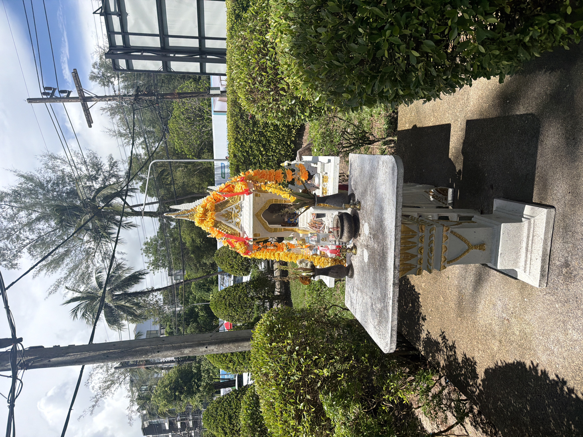 An outdoor spirit house on a concrete pedestal is situated in Choeng Thale, Thailand. The spirit house is adorned with orange marigold garlands and has a white structure with gold embellishments. On the pedestal in front of the spirit house are several items including a small black vase with dried flowers, a small red candle, and a dark bowl containing ash or incense. Behind the spirit house, manicured green bushes form a hedge. In the background, there are palm trees, electrical poles with numerous wires, and buildings. The ground is a mix of concrete and gravel. The lighting suggests it is daytime, with shadows cast by the objects and trees.
