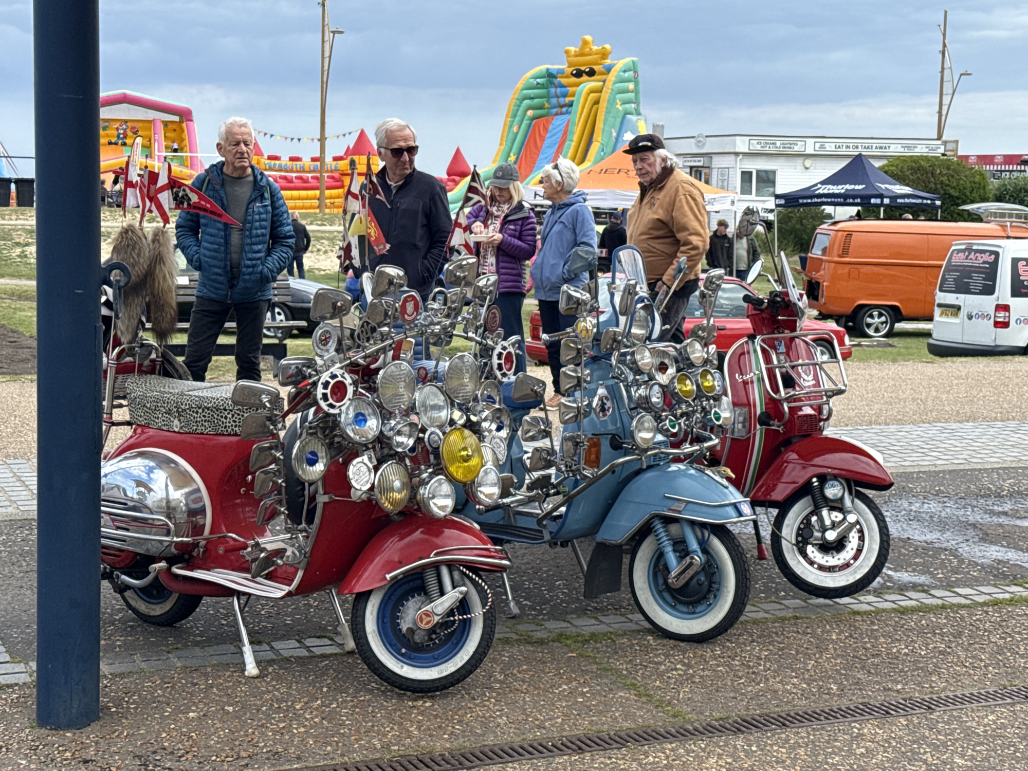 The image captures a scene in Great Yarmouth, United Kingdom, showcasing three highly customized scooters prominently adorned with numerous lights and accessories, indicative of the "Mod" subculture. The scooters are arranged in a row on a paved area, with the closest one being red, the middle one light blue, and the farthest also red.
Several people are visible near the scooters. An older man in a blue jacket stands near the left-most scooter, and a group of four individuals, two men and two women, are near the center, seemingly admiring the vehicles. An older man with a cap is near the light blue scooter.
In the background, an orange van and a white van with branding are parked. Also visible are inflatables in the distance, suggesting a fairground or similar attraction. A building with "Eat In or Take Away" signage is also in the backdrop, along with a blue and white tent. The sky is overcast, indicating potentially dull weather conditions.