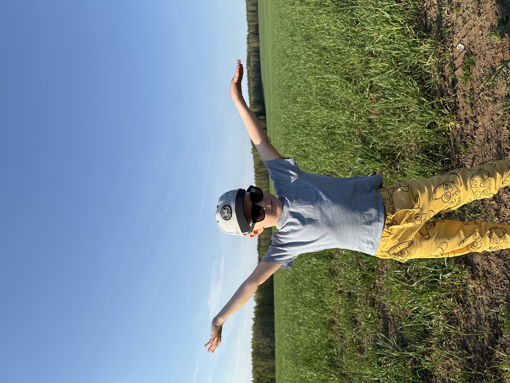 A young boy lies on his back in a field of green grass near Шумиха, Russia. He is wearing a gray t-shirt, yellow pants with cartoon controller patterns, sunglasses, and a gray baseball cap. His arms are outstretched, almost perpendicular to his body. The sky is a clear blue. In the background is a line of trees. The dirt on the far right of the photo is the edge of the field.