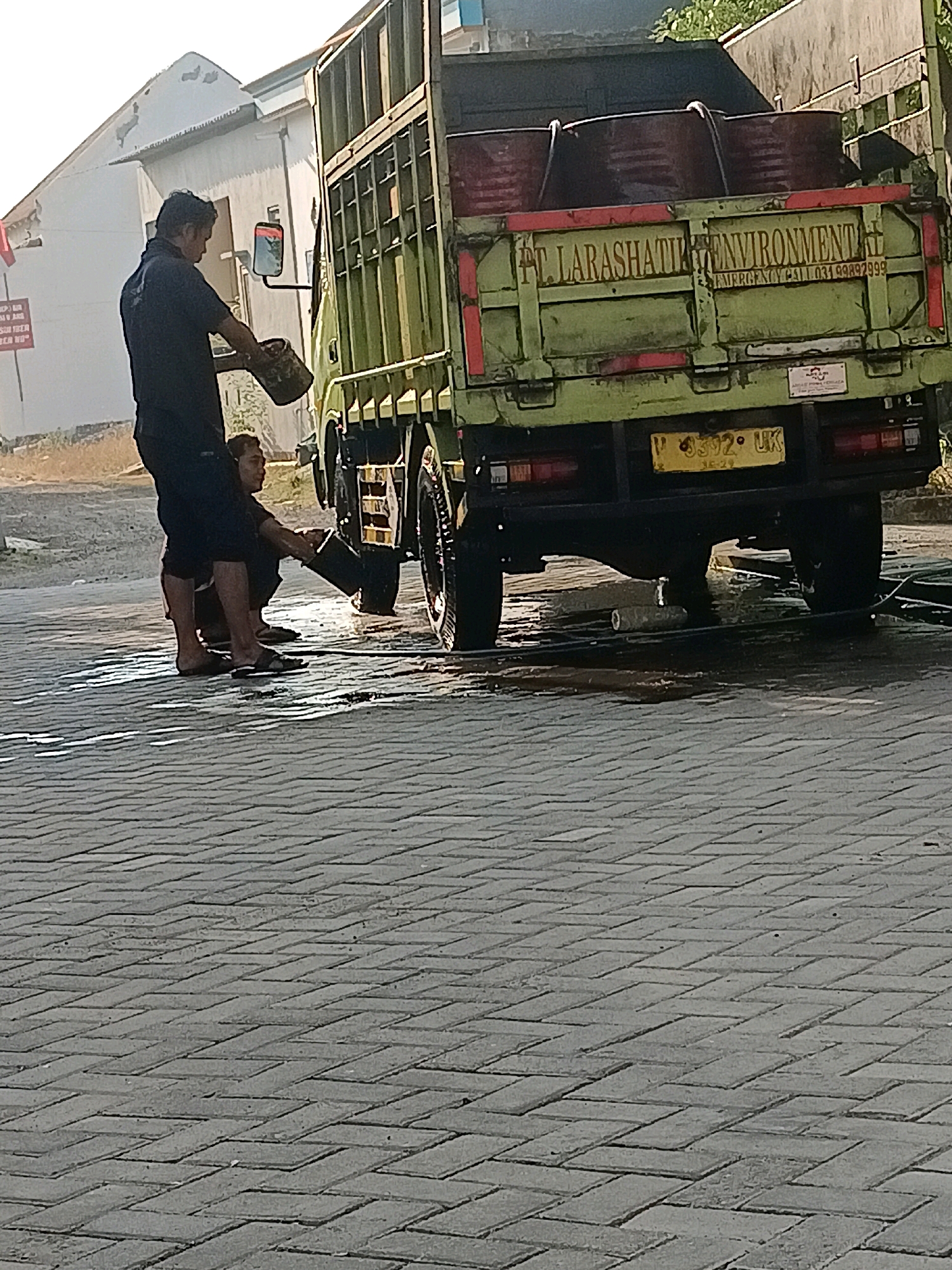 In the city of Krembangan, Indonesia, on a paved street with herringbone brickwork, two individuals are engaged in an activity next to a green truck. The truck has "PT. LARASHATIK ENVIRONMENTAL" and an emergency phone number printed on its side. The truck also has a yellow license plate reading "P 6392 UK." One person, standing and wearing dark clothing, is holding a bucket and appears to be pouring liquid from it. The second person is squatting beside the truck, reaching towards the rear wheel, possibly interacting with the truck's undercarriage. Water is visible on the ground around them, suggesting a washing or cleaning process. The background includes a white building with a visible red sign and some greenery. The lighting indicates it is daytime.