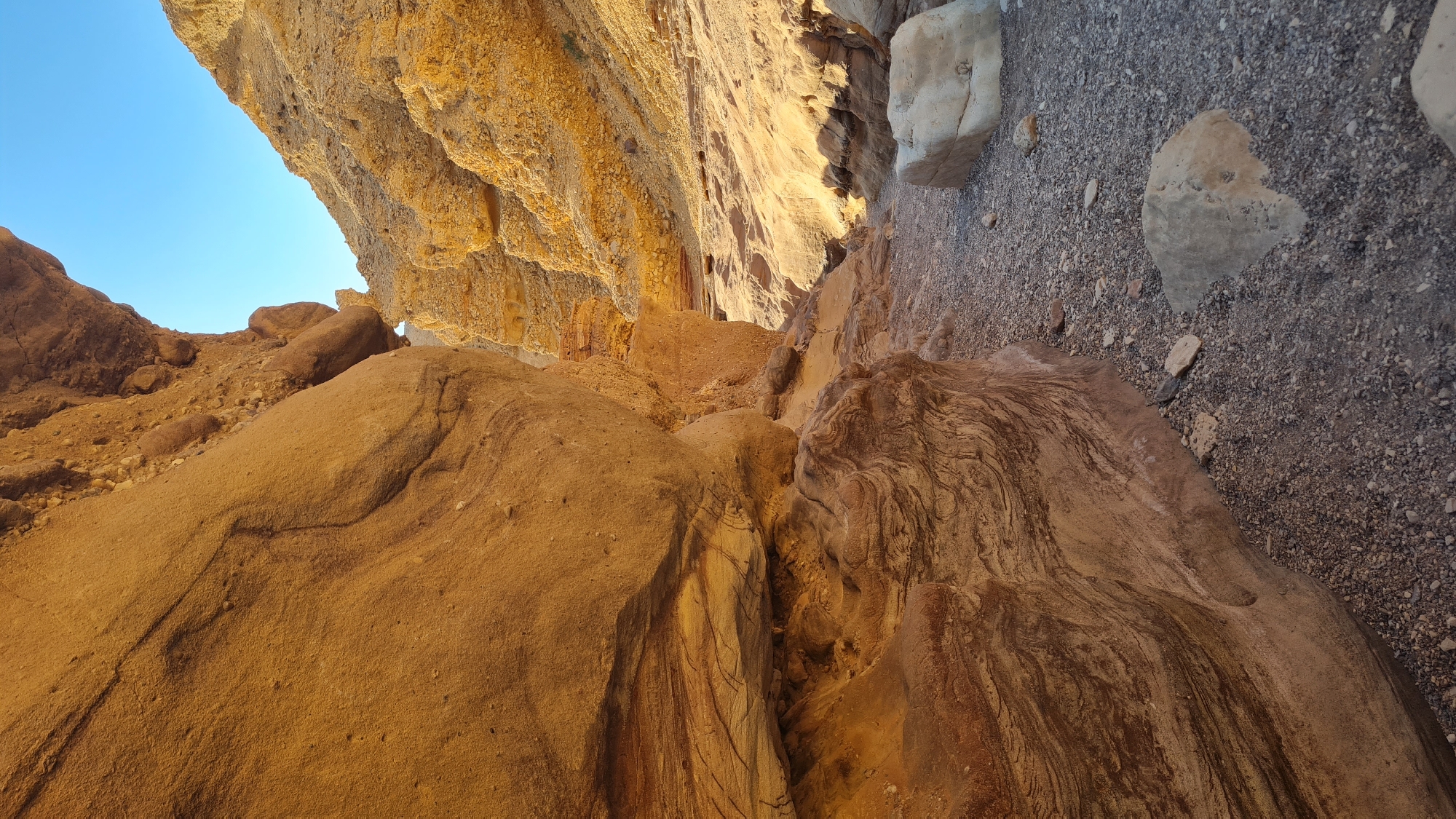 The image depicts a geological formation in Hevel Eilot Regional Council, Israel, showcasing layers of sedimentary rock. Predominantly brown and tan hues characterize the rock surfaces, revealing diverse textures. The rock formation appears rugged, with an uneven and slightly angled orientation. A patch of blue sky is visible in the upper left corner. The composition and setting imply an arid or desert environment.