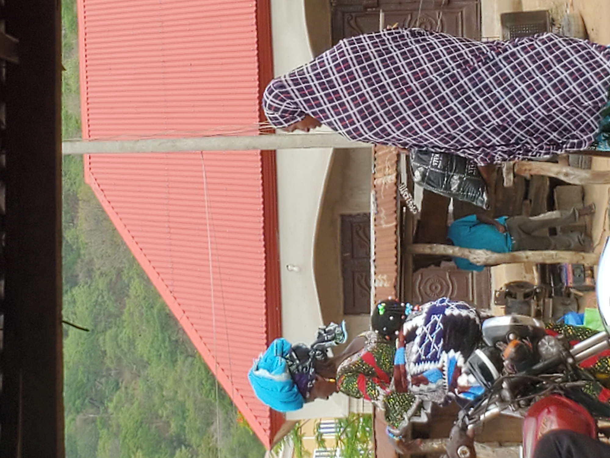 Two people, one wearing a checkered head covering and the other a turquoise head covering, are near a red roof. The scene includes a building with brown doors and trees in the background. It appears to be a residential area. While the precise location is not verifiable based solely on the image, it aligns with scenes common in Mararaba, Nigeria.