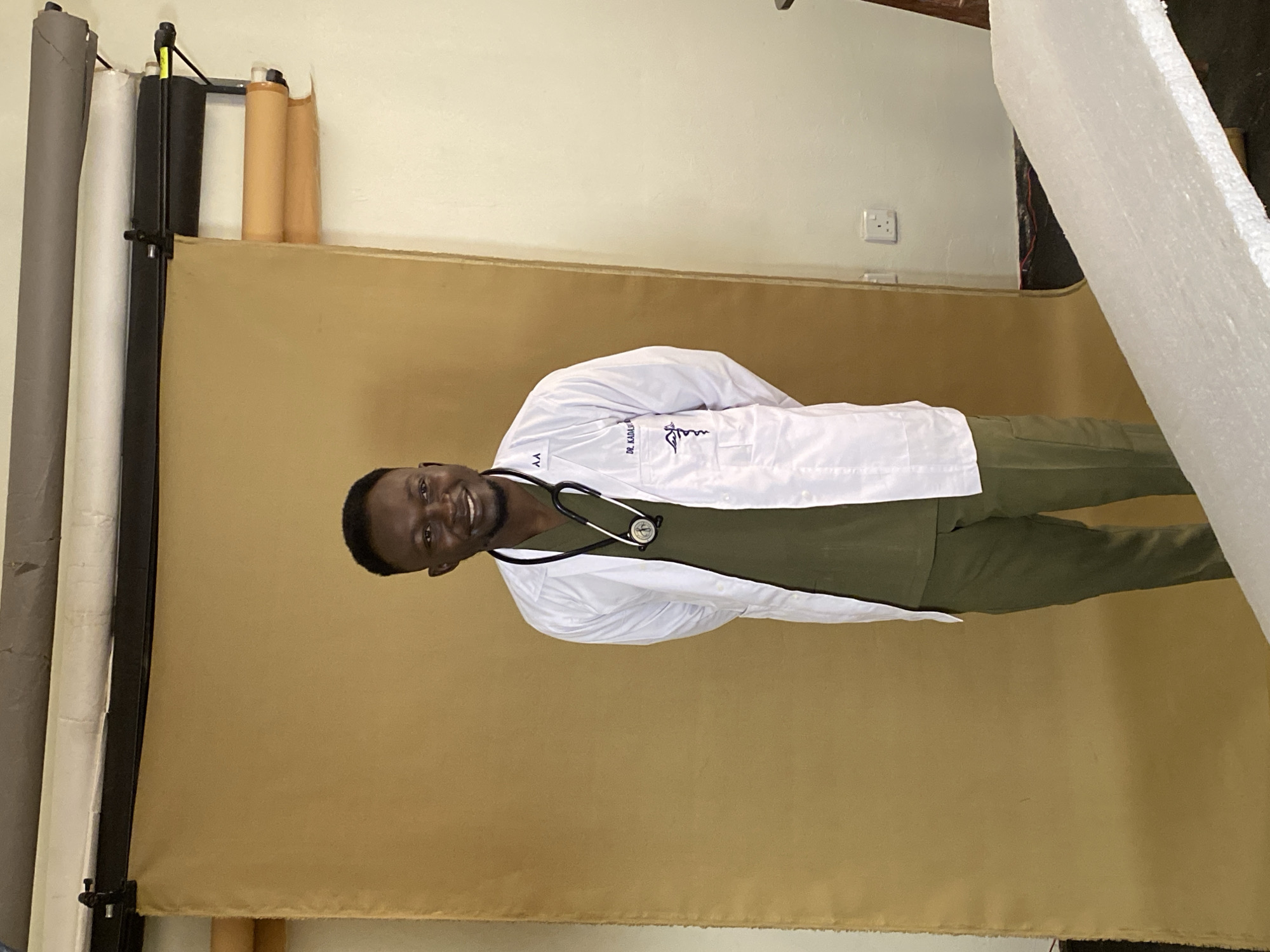 A man in a white lab coat and green scrubs stands in front of a tan backdrop. He has a stethoscope around his neck and is smiling at the camera. There is a name tag on his lab coat that reads "DR. KADAS. J." The man is positioned in the center of the frame, with his hands clasped behind his back. The background suggests an indoor studio setting, possibly a photography studio or a room prepared for formal portraits. The lighting appears to be artificial, casting soft shadows. The overall impression is of a professional portrait. The location is identified as Jos, Nigeria.