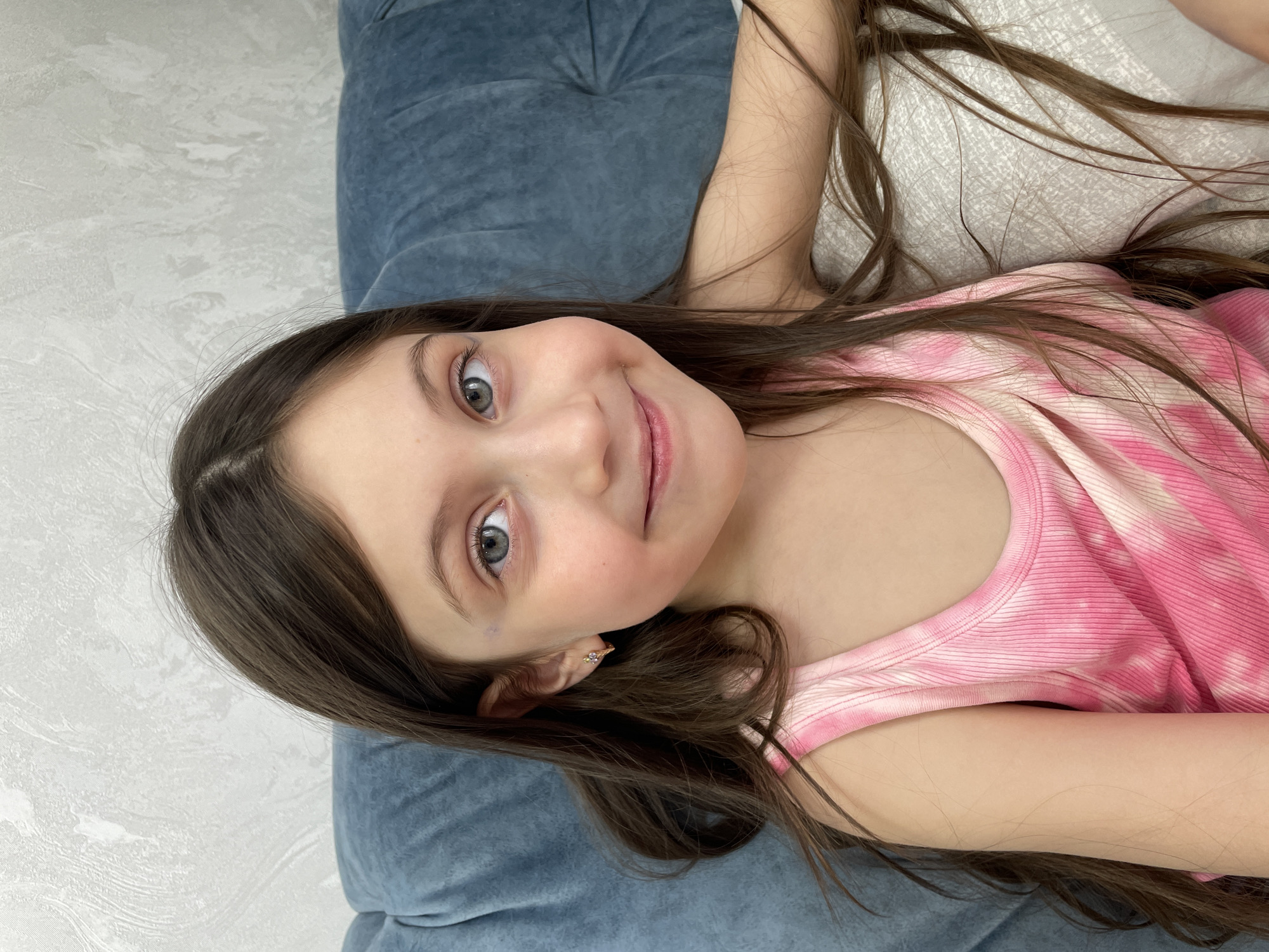 A young girl with dark hair is lying down on a light gray patterned surface next to a dark blue cushion. She is wearing a pink tank top. Her face is turned towards the camera with a slight smile. She has light eyes. She is wearing a small earring in her left ear. Her hair is long and spread around her head. The exact location in the city of Izhevsk, Russia, cannot be determined from the image.
