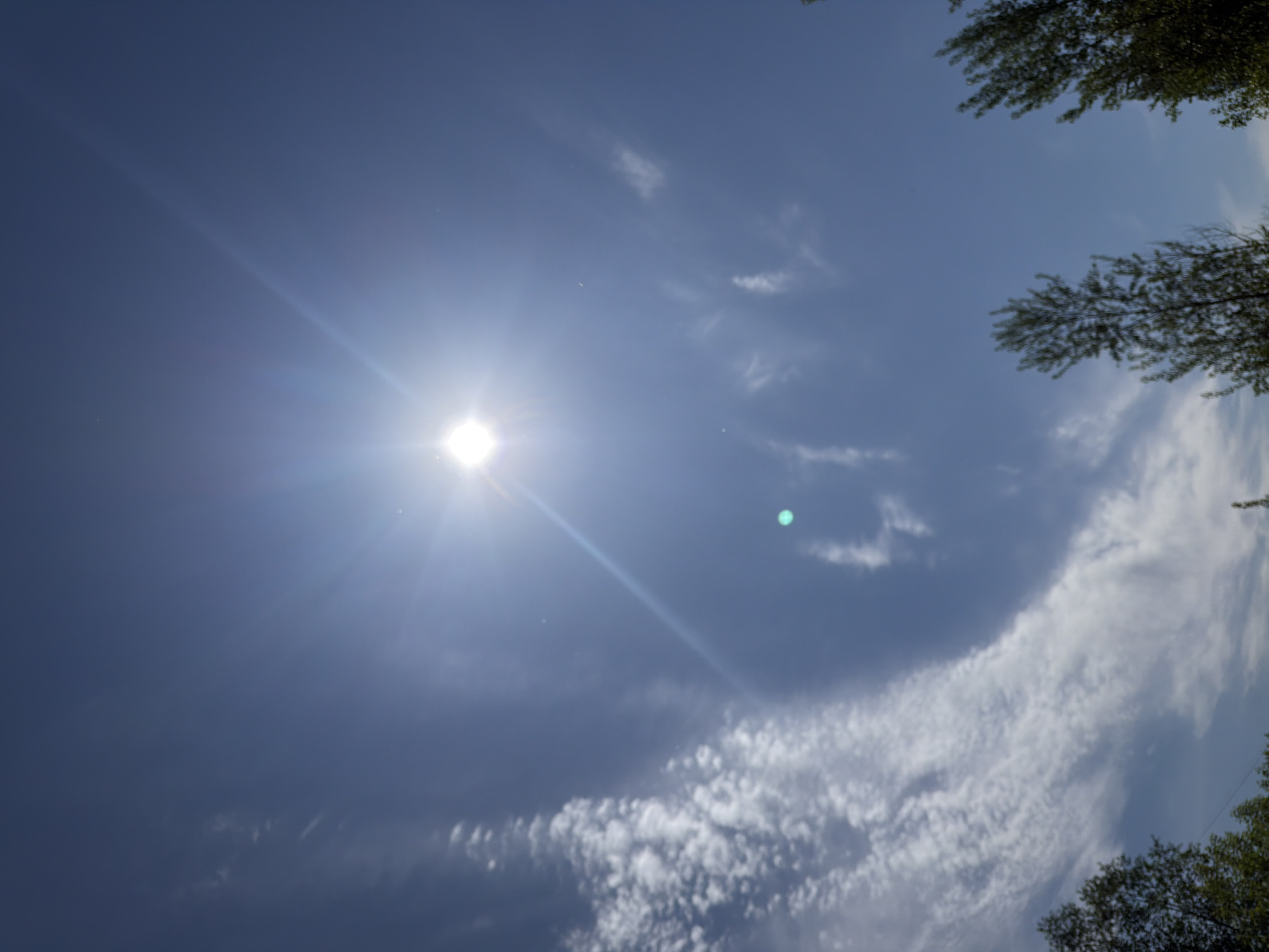 The image depicts a daytime scene in Chaikovsky, Russia, characterized by a bright sun in a blue sky with scattered clouds. The sun is positioned prominently in the upper left quadrant of the frame, emitting strong rays of light. A notable feature is a swirling cloud formation in the lower right quadrant, composed of numerous small, white cloudlets. Some treetops are visible along the right edge of the frame.