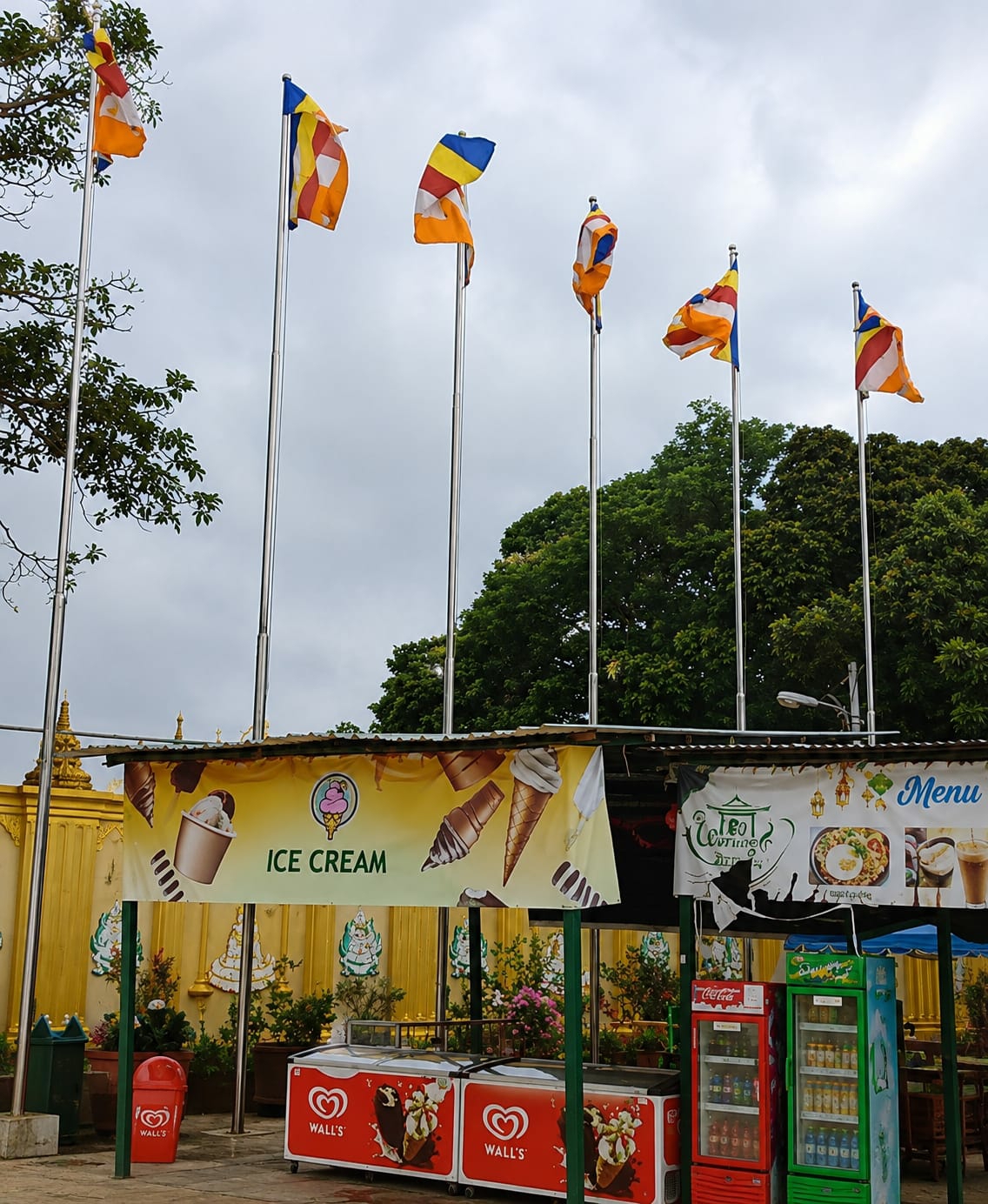 In the city of Dolat Rayat, Indonesia, a street-side vendor stall is depicted under a cloudy sky. Multiple Buddhist flags fly from tall poles above the stall. The stall itself features a yellow banner advertising "ICE CREAM" with images of ice cream cones and cups. To the right of the ice cream banner, another banner displays food items and the word "Menu." Below the banners, two large, red and white "Wall's" branded ice cream freezers are visible, along with two smaller refrigerators, one red and one green, stocked with drinks. Red and green trash bins are placed near the front of the stall. The background shows a golden wall with decorative carvings and lush green trees. No people are clearly visible in the image.