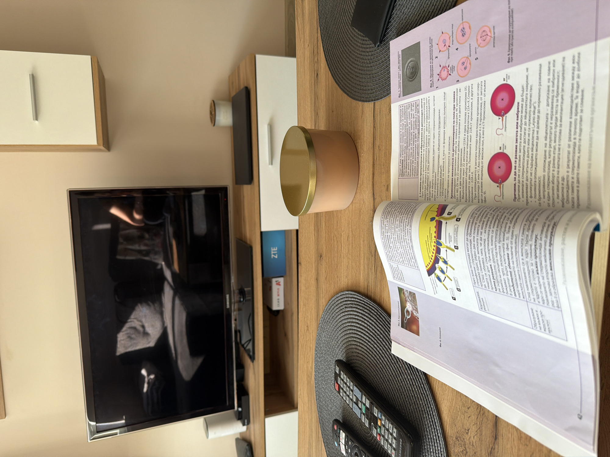 The scene is an indoor shot of a wooden table, likely in Litakovo, Bulgaria. On the table, there's an open biology textbook with diagrams of human cells and fertilization processes. A beige and gold candle sits near the book. Two gray placemats are positioned on the table, with a black TV remote control on one. Part of a television is visible in the background, along with a beige wall and a wooden shelf. The environment suggests a domestic setting, possibly a living room or study area.