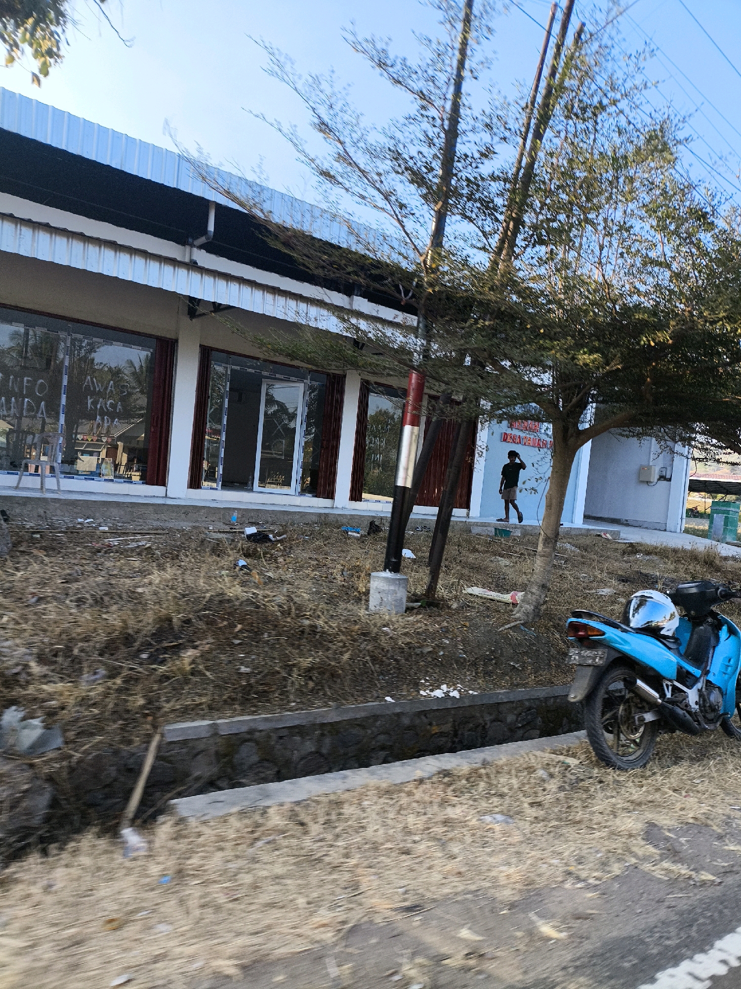A bright blue motorcycle is parked on a dry, grassy area next to a road in Dulupi, Indonesia. The motorcycle, with a white and blue helmet on the seat, is facing left. Behind the motorcycle, there is a concrete drainage ditch filled with stones. Further back, there is a building with large glass windows, some of which have writing on them in white. A person in a dark shirt and shorts is walking towards the right side of the building entrance. There are trees with sparse leaves and branches in front of the building. The sky is clear and blue. The ground in front of the building and along the road is dry and covered in dead grass and some scattered debris.