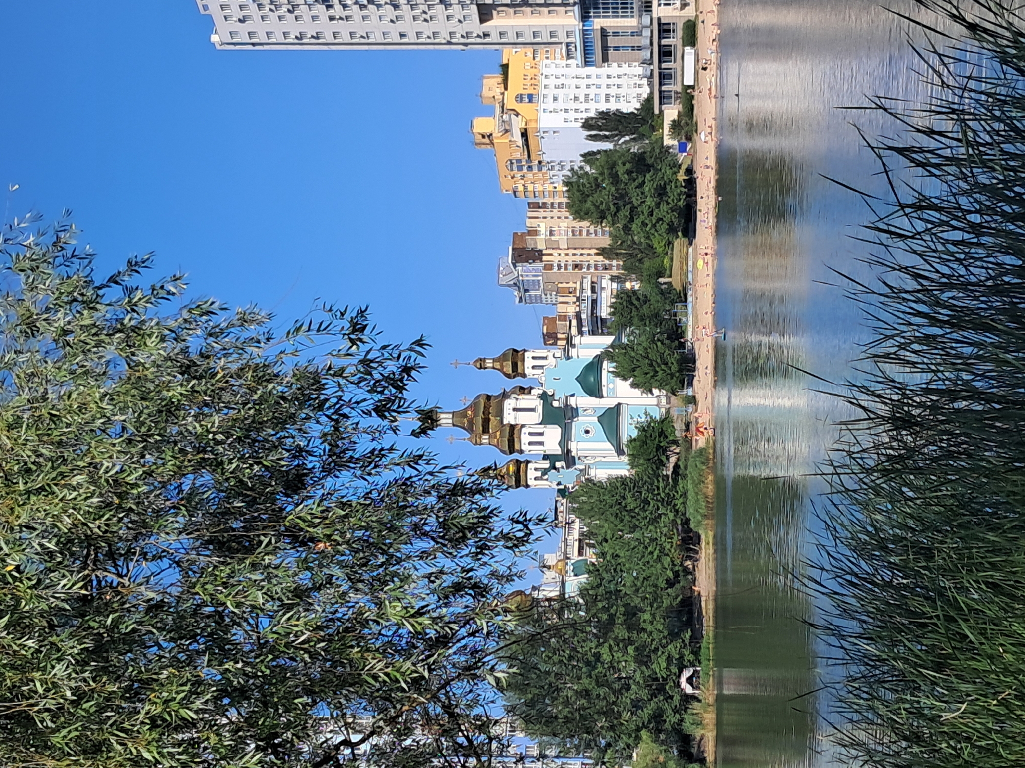 The image captures a scenic view of the Hydropark in Kyiv, Ukraine, on a clear day. In the foreground, tall, green grass sways gently, bordering a tranquil body of water that reflects the sky and surrounding elements. Across the water, a picturesque scene unfolds: a bright blue and white church with golden domes stands out against a backdrop of modern apartment buildings. The church is surrounded by lush green trees, adding a touch of nature to the urban landscape. On the sandy beach across the lake, numerous people are seen enjoying the sunny weather, with some sunbathing, others walking, and a few engaging in water activities. The overall atmosphere suggests a lively summer day at this popular recreational spot in Kyiv.