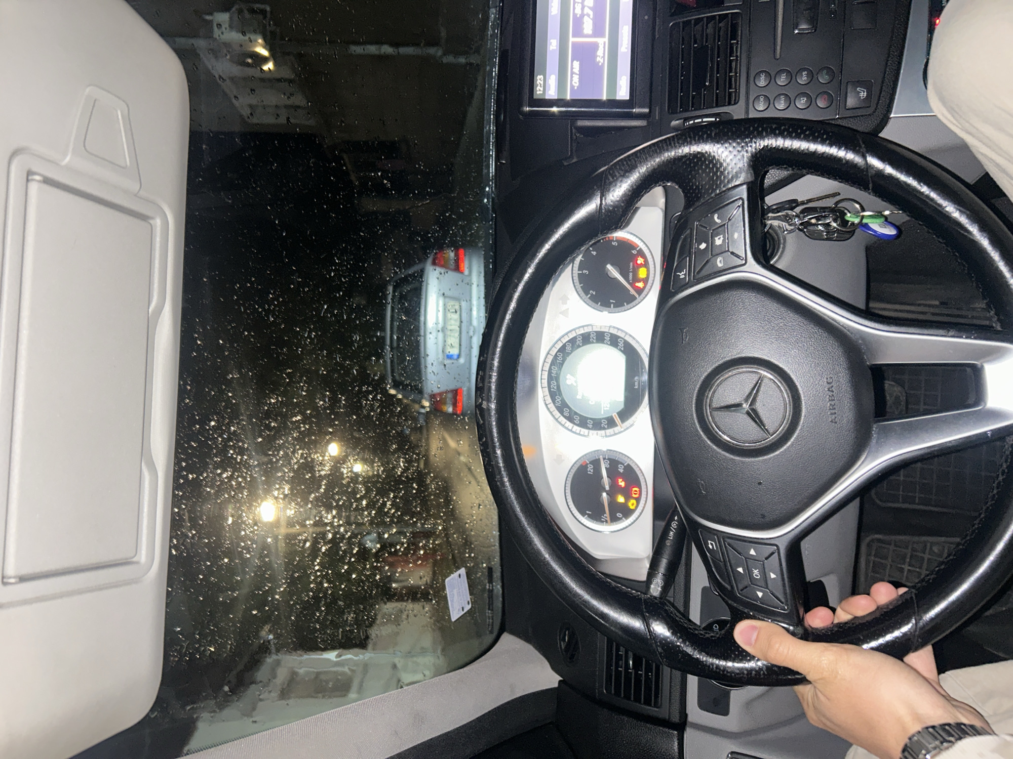 The image shows the interior of a car, likely a Mercedes-Benz, as indicated by the steering wheel and dashboard design. It appears to be nighttime or a dimly lit environment, and it's raining, as seen through the windshield. A car is visible in front, through the windshield. The driver's hand is visible on the steering wheel, and a wristwatch can be seen on the wrist. The radio screen is lit, showing a radio station. The license plate is partially visible on the car in front. Several dashboard indicator lights are illuminated. The overall impression is that the person is driving in rainy conditions.