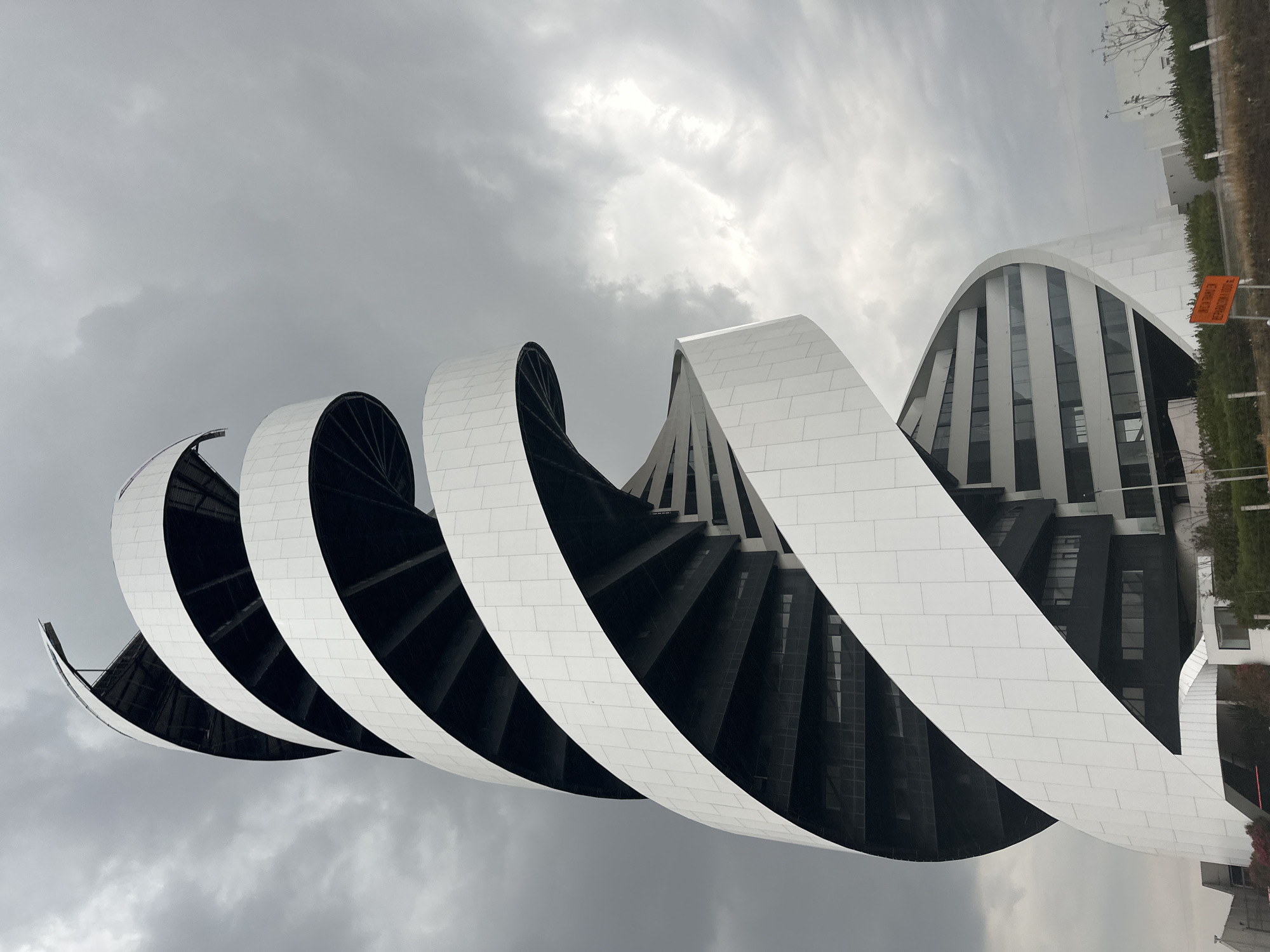 The image depicts a modern architectural structure in San Bernardino Tlaxcalancingo, Mexico, under a cloudy sky. The building features a spiral design with alternating white and black sections, creating a visually striking contrast. The white sections appear to be composed of tiled or modular panels. The black sections reveal the building's inner structure with vertical windows. In the background, there is minimal vegetation, along with other buildings and a highway.