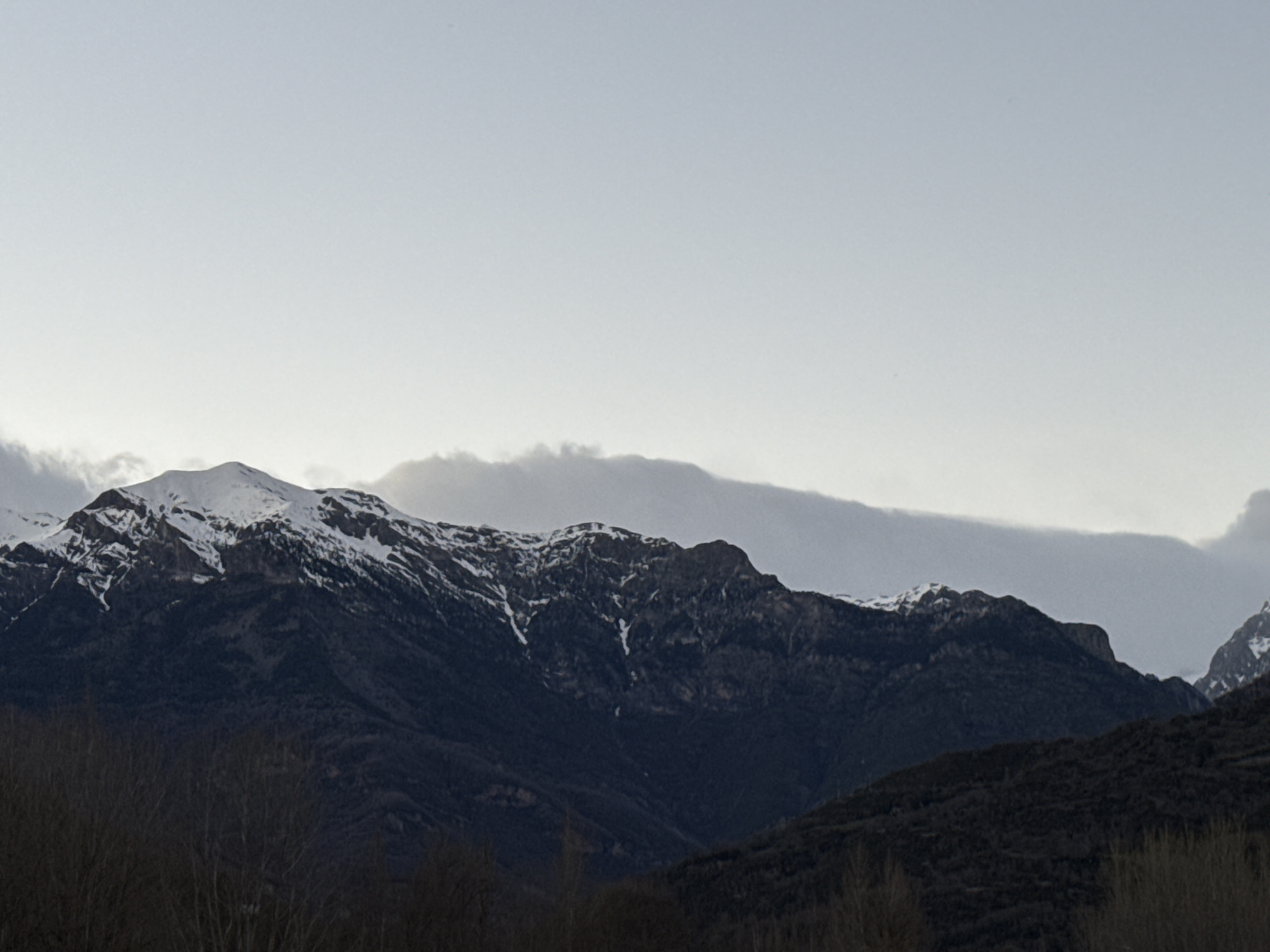 The image shows a mountain range partially covered in snow, likely taken near Castejón de Sos, Spain. The sky is overcast and gray, providing soft, diffused light. In the foreground, there are dark, leafless trees, indicating either a winter or early spring season. The mountains are rugged with visible rocky outcrops and patches of snow clinging to the higher elevations. The overall tone is muted and somber.