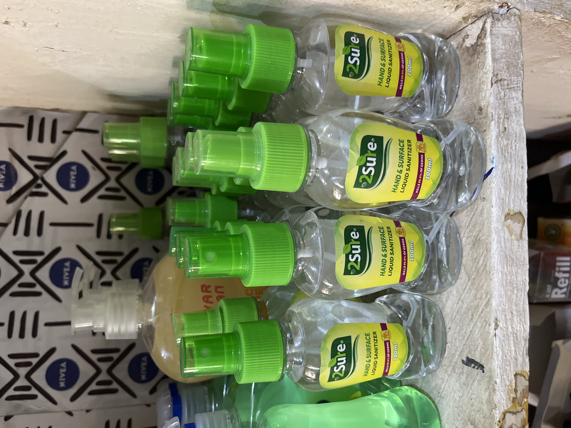 In Bukuru, Nigeria, a shelf holds several bottles of 2Sure Hand & Surface Liquid Sanitizer. The bottles have clear plastic bodies, green spray caps, and yellow labels detailing the product's name, purpose, and that it kills 99.9% of germs and contains 70% alcohol. The shelf itself is worn and appears to be made of wood. In the background, a patterned fabric with "NIVEA" logos is visible. Additional green-capped bottles and other products are partially visible on the shelf and behind the main subjects.