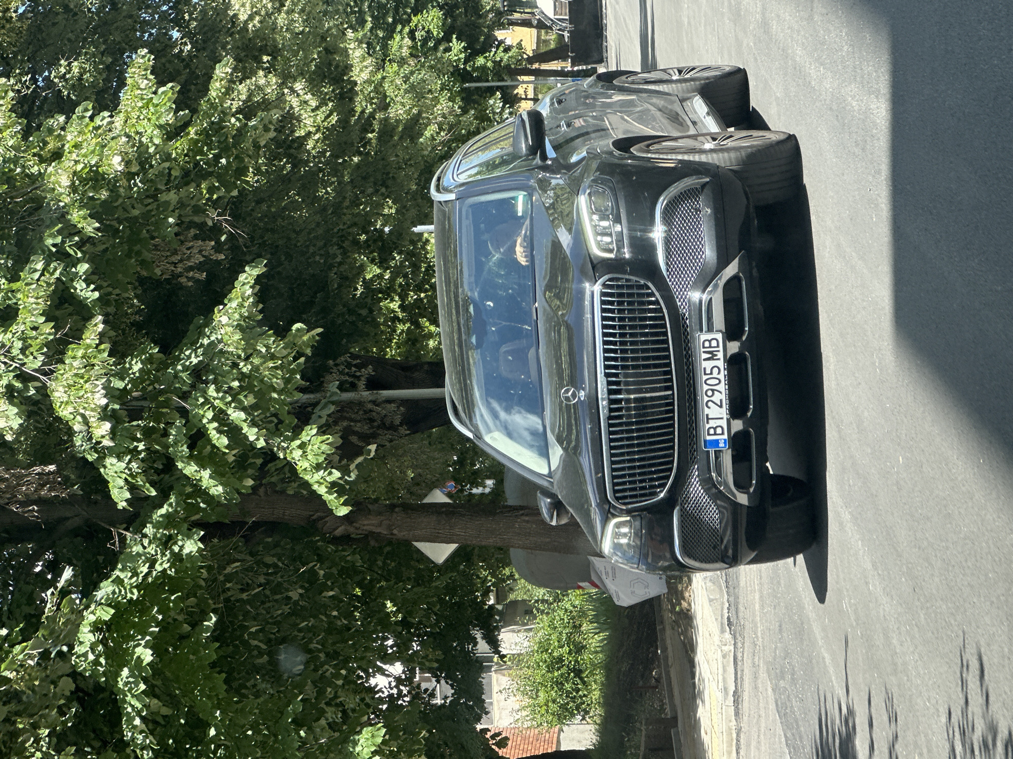 A dark colored, luxury SUV, possibly a Mercedes-Benz Maybach GLS, is driving down an asphalt road lined with trees and buildings. The vehicle has a large grille with vertical chrome bars and the Mercedes-Benz logo at the top. The license plate reads "BT 2905 MB". The driver, a person with light-colored skin and short dark hair, is visible through the windshield. Sunlight filters through the dense green foliage of the trees, casting shadows on the road. In the background, a building with a red brick facade and some bushes are visible. The scene appears to be in an urban environment on a sunny day.
