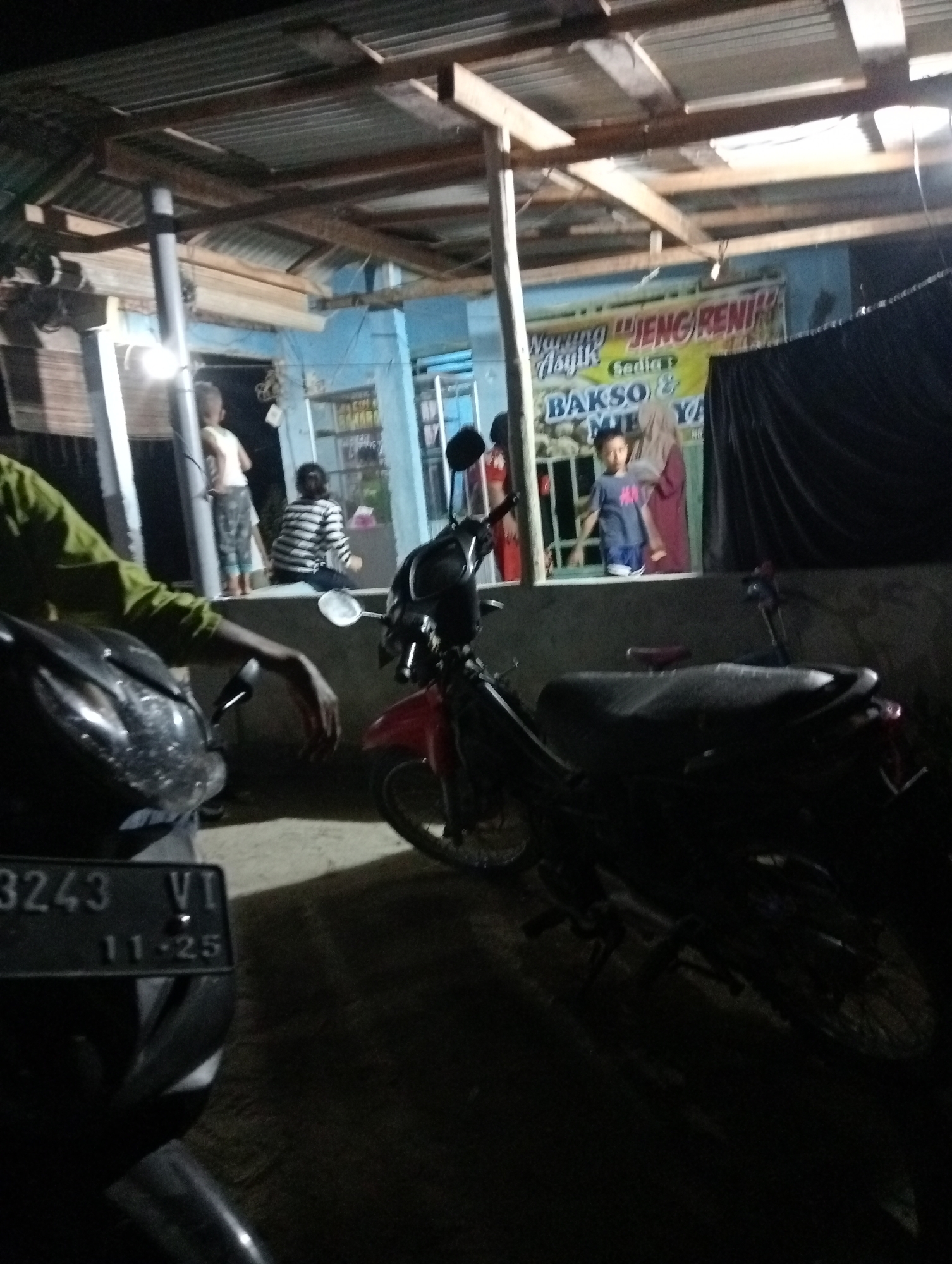 The image depicts a nighttime scene outside a food stall in Rasuan Baru, Indonesia. A handwritten sign in Indonesian, partially visible, advertises "Warung Asyik Meng Reni" and "Bakso & Mie Ya". In the foreground, a motorcycle is parked, and another motorcycle is partially visible to the right. Two children are standing near the counter of the food stall, and an adult wearing a hijab is also present. Another individual, possibly an adult, is seen from the back, wearing a striped shirt. The environment appears to be an outdoor setting with a corrugated metal roof structure over the food stall. The overall lighting suggests artificial illumination from the stall.