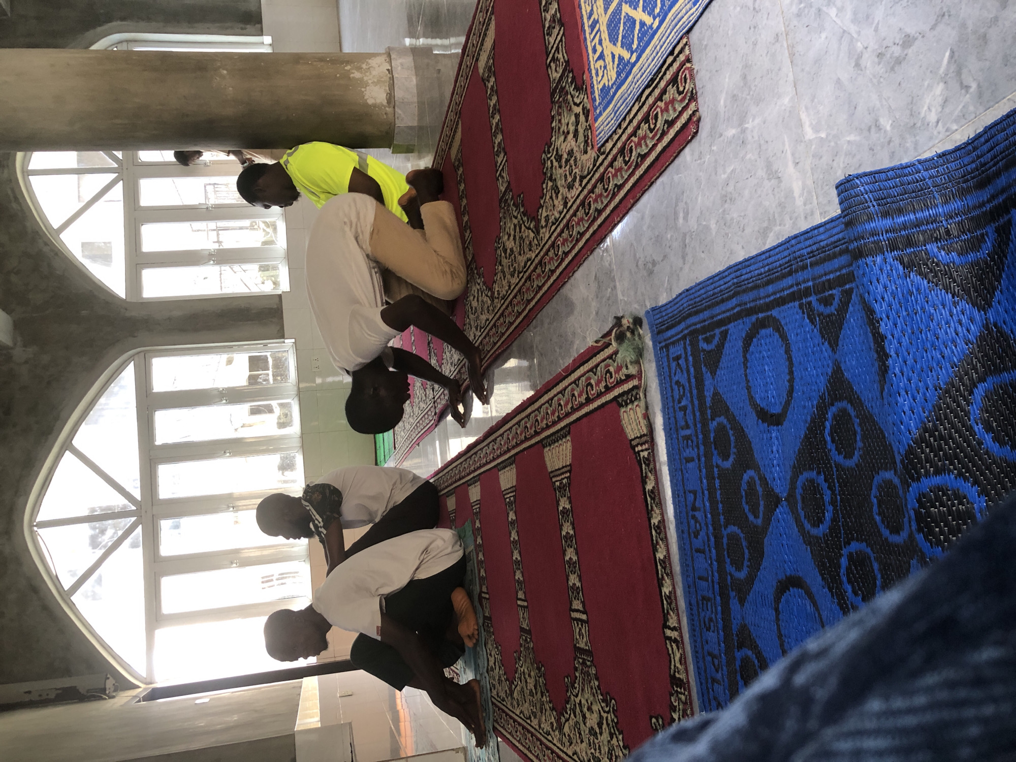 In the city of Unknown, Unknown, a mosque interior is depicted with several individuals engaged in prayer. The scene is characterized by large arched windows with multiple panes that allow abundant natural light to illuminate the space. The floor is tiled with a marble pattern, and prayer mats are laid out on the floor. In the foreground, a person's legs are visible, covered by dark blue denim, suggesting they are seated or kneeling on a blue woven mat. Further back, four men are the focus of the image. One man, wearing a white t-shirt and dark pants, is in a kneeling position, leaning forward with his hands on the floor, mid-prayer. Another man in a white t-shirt and tan pants is in a similar prayer posture. To the right, a man in a bright yellow, high-visibility shirt is standing, facing away from the camera. Another man, partially obscured, is also in prayer behind him. The walls appear to be a light neutral color, with decorative arched window frames. A large, round pillar is also visible, contributing to the architectural style of the mosque. The lighting suggests it is daytime, with the sun casting bright light through the windows.