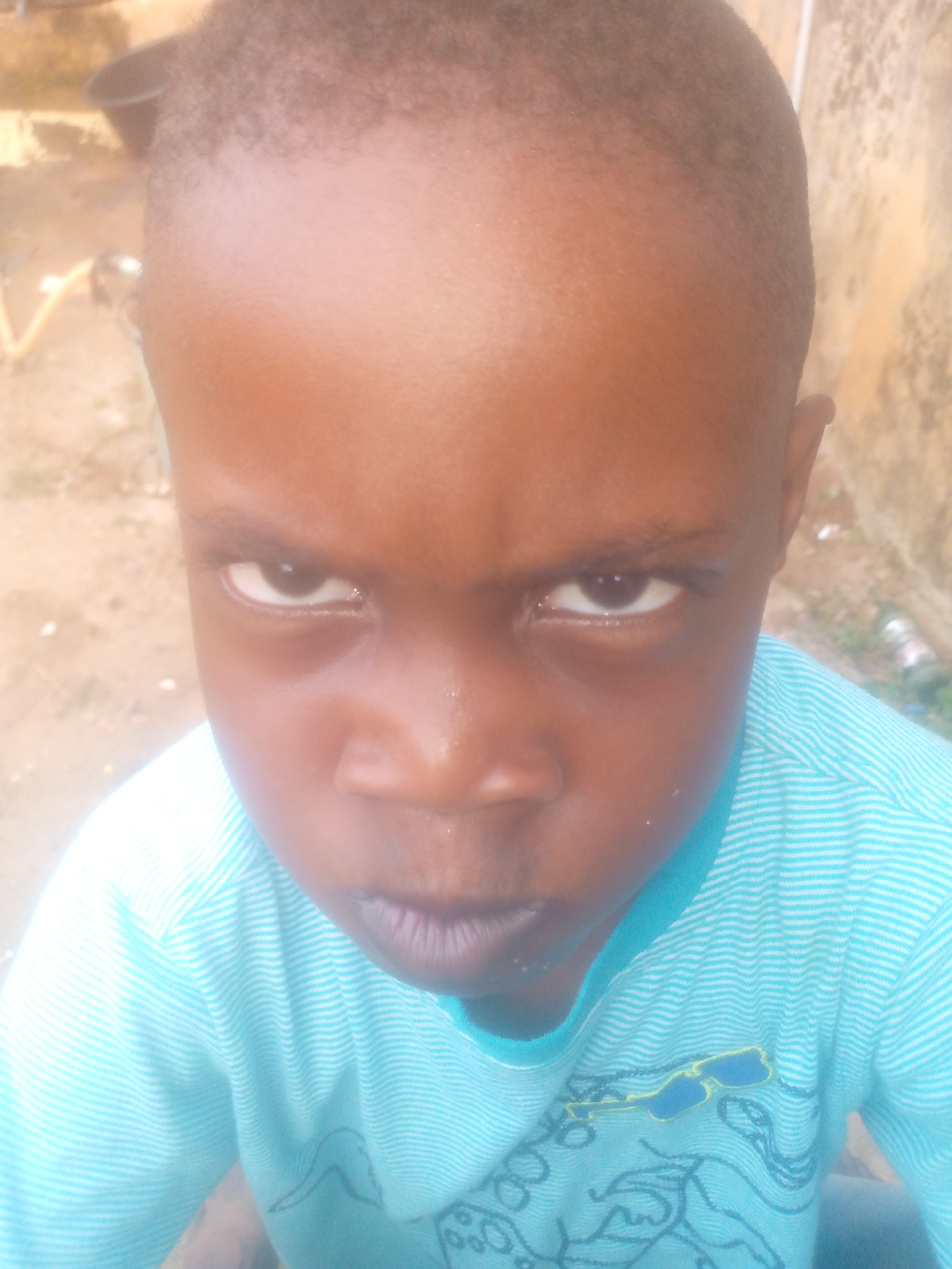 A close-up shot shows a young boy with dark skin, staring intently at the camera with a slightly furrowed brow. He is wearing a blue and white striped long-sleeved shirt with a printed design. The background is blurred, showing a wall and some foliage. The location details are not available.