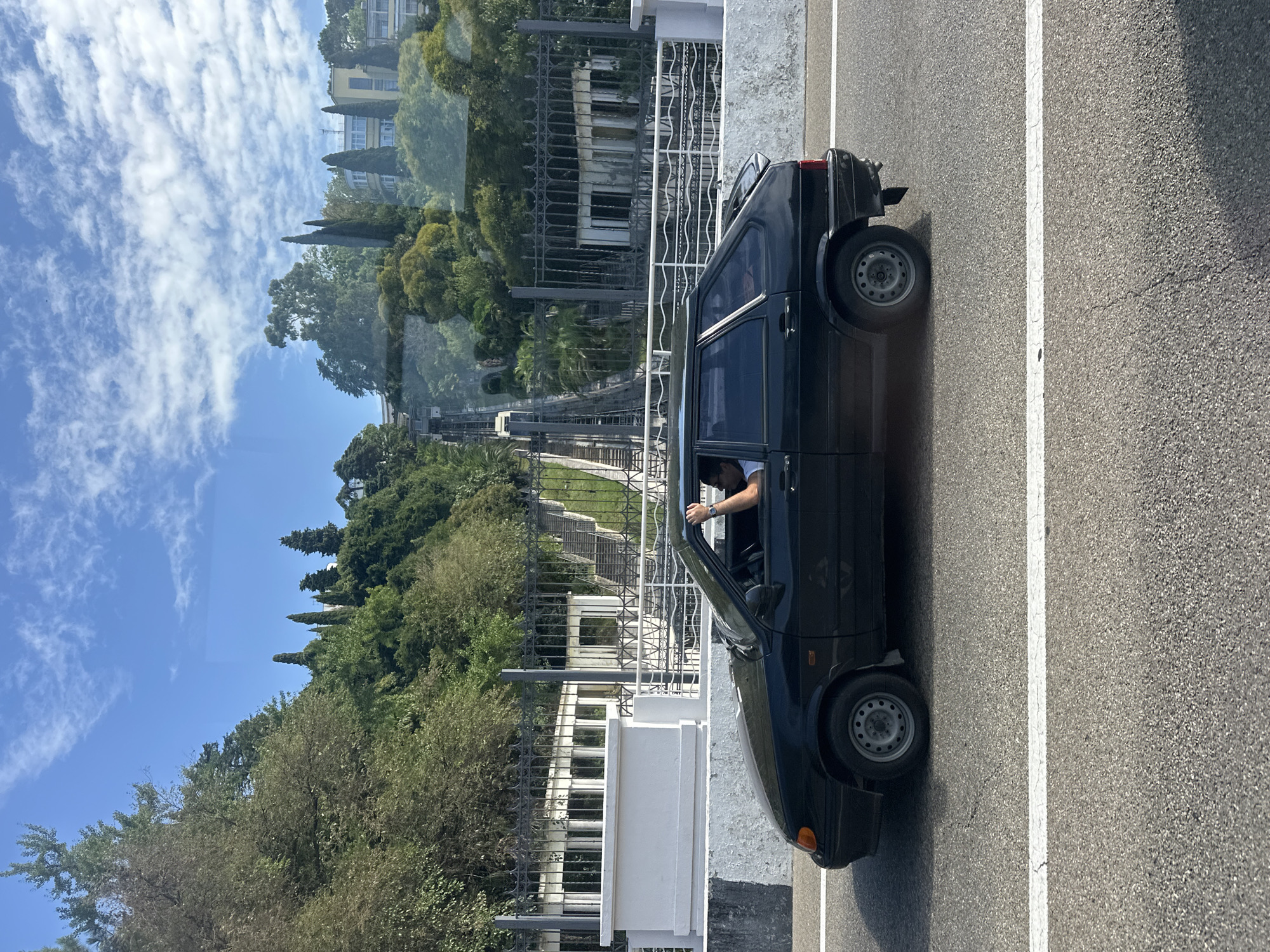 This image captures a view of a black hatchback car driving on a paved road with white lane markings. The car is viewed from the side, showing its left side and the driver inside. The driver, a male, is visible with his left arm raised and hand on the top of the car door frame. He is wearing a watch on his left wrist. Behind the car, a hillside is covered with dense green trees and vegetation. A black metal fence runs along the hillside, behind which is a white structure with columns and a set of stairs leading upwards. Further up the hill, there are buildings and more trees. The sky above is blue with scattered white clouds. This scene is consistent with the landscape found in Sochi, Russia, potentially near an area with a funicular or inclined railway, suggested by the parallel tracks visible on the hillside in the background. The overall environment appears to be a sunny day in a city known for its mountainous terrain and lush greenery.