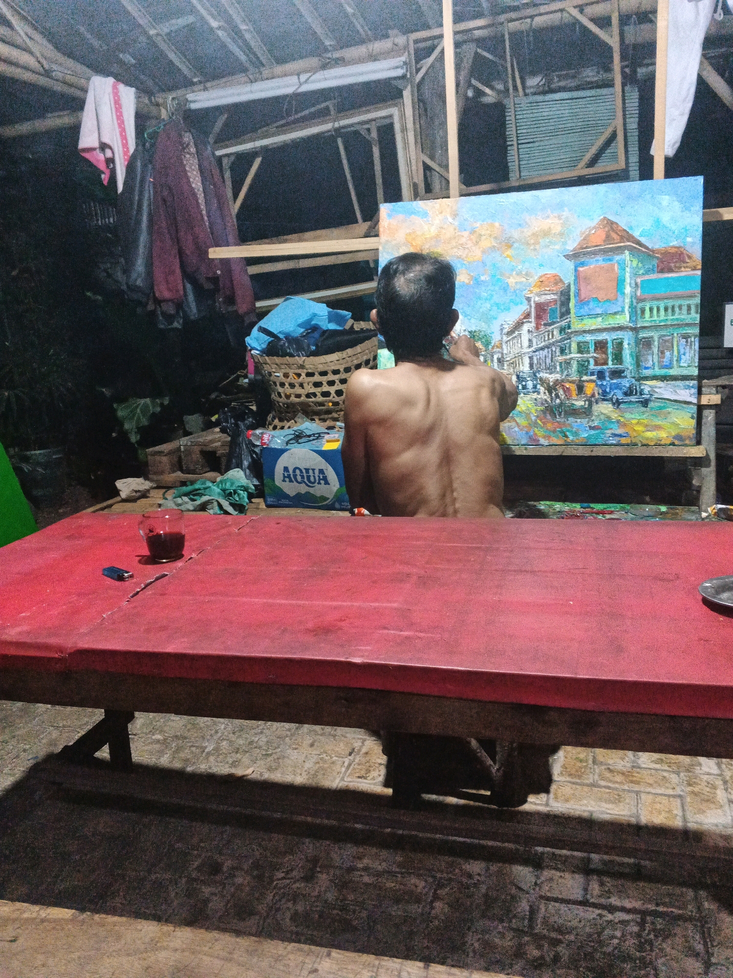 An adult male, shirtless and with a visible spine, is painting a vibrant canvas depicting a street scene with buildings and vehicles. He is positioned in a rustic, outdoor setting, possibly a workshop or an open-air studio, characterized by a bamboo-and-wood roof and exposed structural elements. In the foreground, a red table displays a glass of dark liquid, a lighter, and other assorted items. Various objects are scattered around the area, including a woven basket containing a blue bag, a bottle of Aqua, and some crumpled green cloth. Clothes hang on a makeshift rack in the background, suggesting a residential or utilitarian space. The overall scene has a slightly dim, possibly evening ambiance. Based on the Aqua bottle, a prominent Indonesian brand, the location is likely Indonesia. While specific details about Ungaran are not directly visible, such as street names or distinct landmarks, the presence of Aqua, a common commodity in Indonesia, and the architectural style in the painting could be consistent with areas in or around Ungaran, a town in Central Java, Indonesia.