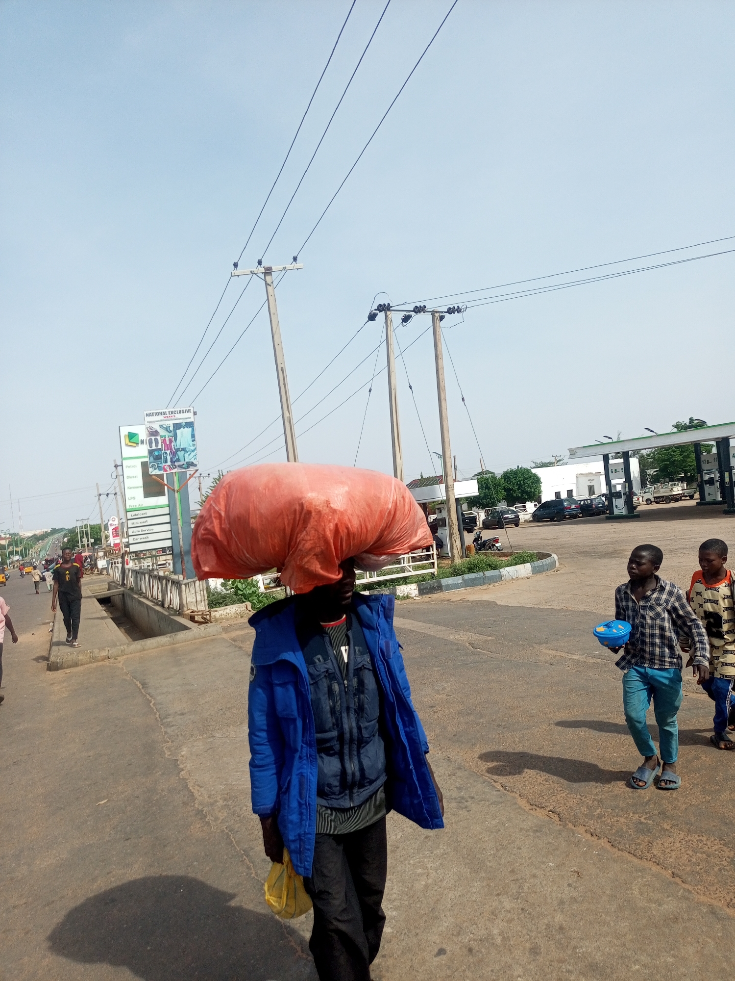 A medium shot captures a street scene in Gombe, Nigeria, on a clear, sunny day. In the foreground, a man walks towards the camera, carrying a large, bright orange bundle balanced on his head. He wears a dark jacket over a patterned shirt and black pants, and holds a yellow bag in his right hand. To his right, two young boys walk away from the camera. The boy on the left wears a plaid shirt and blue pants, holding a blue bowl. The boy on the right wears a patterned shirt and dark pants. In the background, a gas station with multiple pumps is visible, with several cars parked. Power lines and utility poles stretch across the sky. To the left, a street sign for "NATIONAL EXCLUSIVE" and a list of fuel types and services is prominent. Other people and vehicles are present on the street and sidewalk, suggesting an active urban environment.
