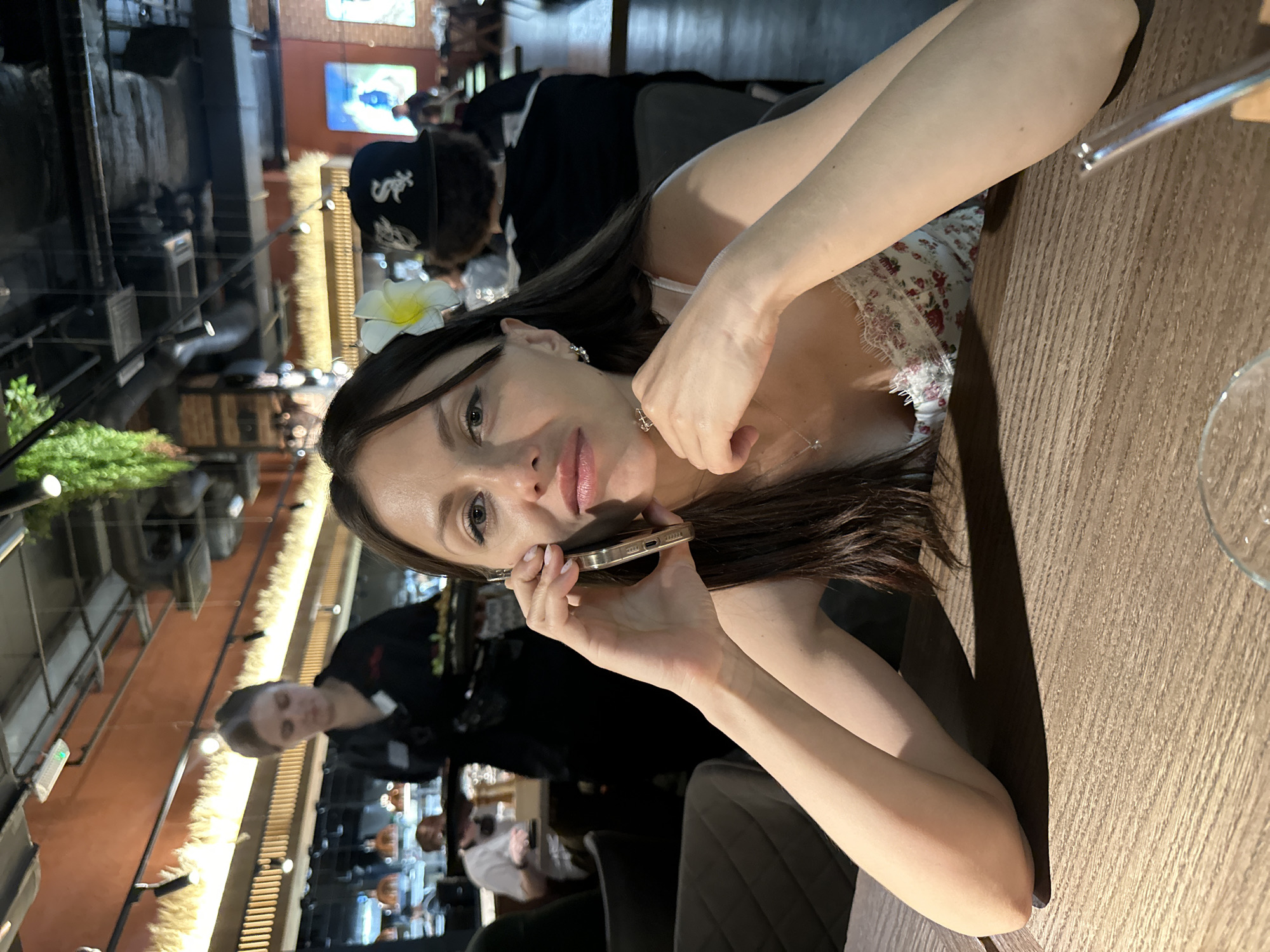 A young woman sits at a wooden table in a restaurant setting, engaged in a phone conversation. She is holding a gold-colored smartphone to her ear with her left hand and has dark hair. She is wearing a light-colored, floral patterned top with a lace trim. The restaurant's interior includes exposed brick, industrial-style lighting, and a dark-colored ceiling with visible ventilation and piping systems. Several other individuals are present, including a person wearing a black baseball cap with white lettering and other seated patrons in the background. A waiter is visible behind the woman. The scene is illuminated by warm-toned lighting, casting shadows across the table and contributing to the ambiance of the restaurant. There is no location information in the image.