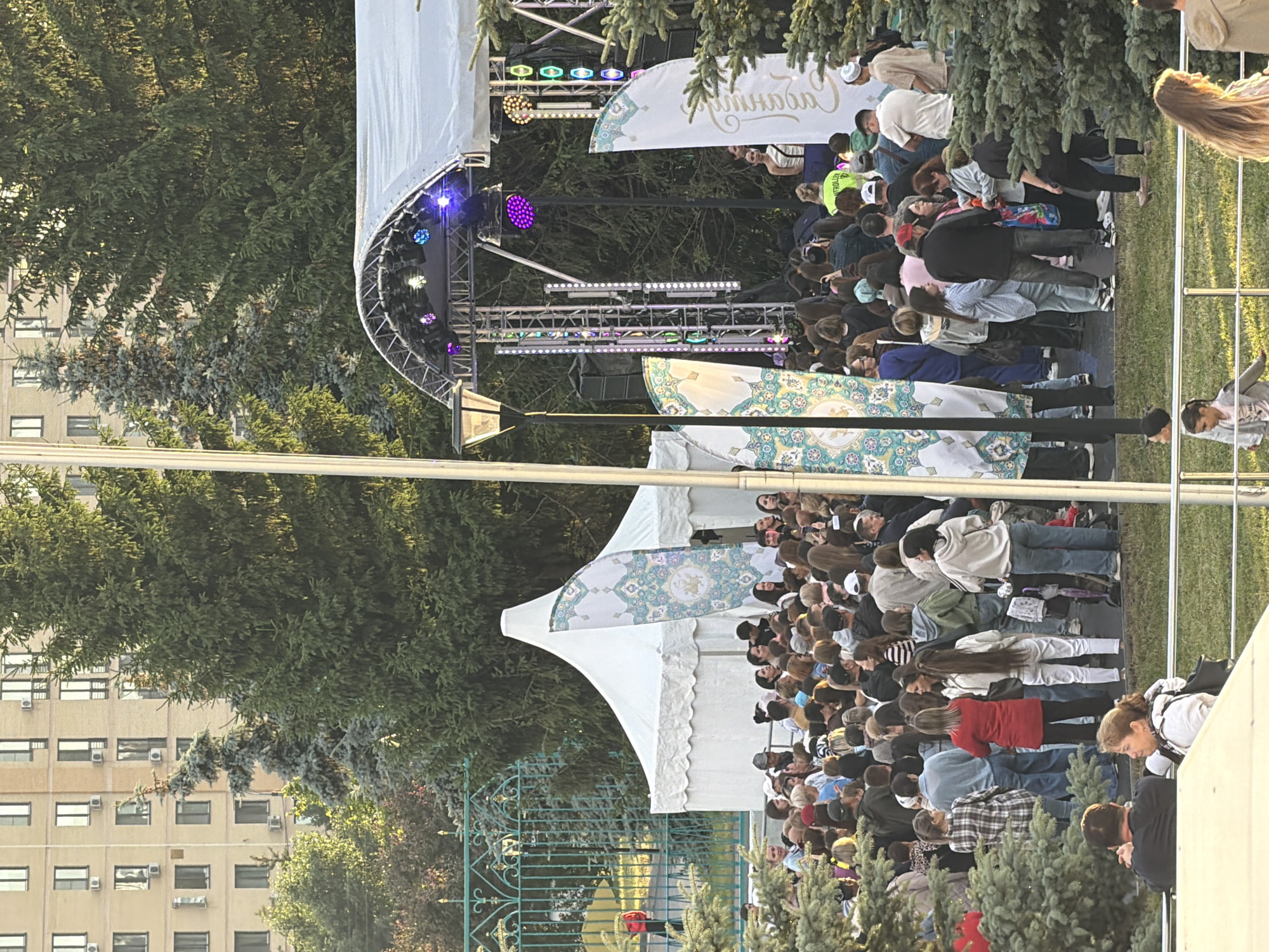 In an outdoor urban setting, a large crowd of people has gathered near a temporary stage and tents. The stage is adorned with lighting, and several banners with intricate patterns are visible. The environment is characterized by lush green trees, suggesting a park or public square. In the background, a multi-story building with numerous windows stands, indicating the presence of an urban landscape. People are standing, watching the stage, or milling about, some holding phones, possibly recording the event. The atmosphere appears to be festive, with a lively gathering in progress.