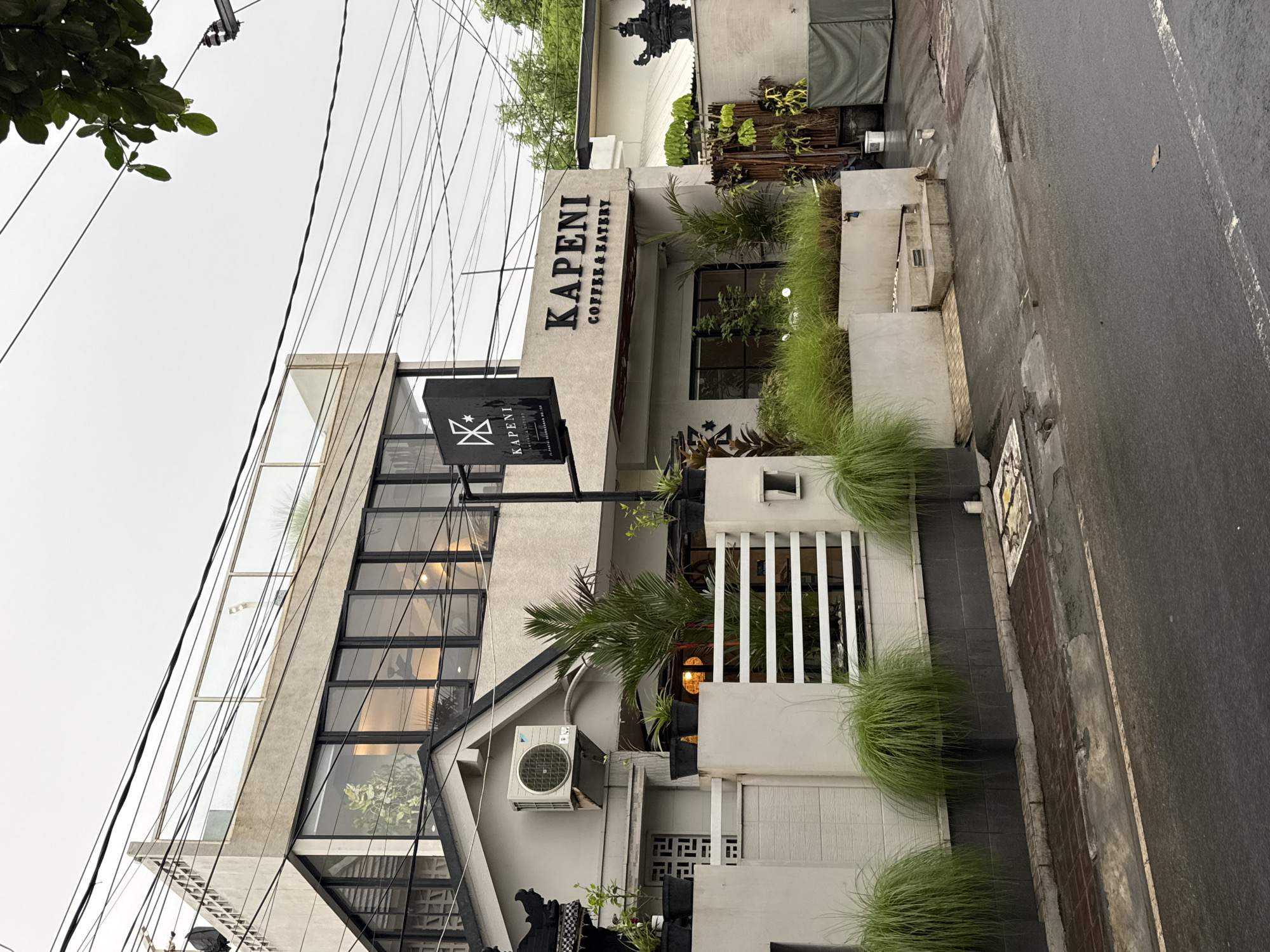 The image presents the exterior of "KAPENI COFFEE & EATERY," a modern multi-story building situated on a street in Kuta, Indonesia, likely during the daytime under an overcast sky. The building features a contemporary design with light grey concrete walls and large, black-framed glass windows.
A prominent black sign with raised white letters on the building's facade clearly displays "KAPENI COFFEE & EATERY." Below this, a black rectangular hanging sign features a white, stylized logo that resembles a crown or a letter 'K', with "KAPENI" written underneath.
The area around the coffee shop is well-maintained with landscaping, including rows of decorative green grasses along the sidewalk and various potted plants near the entrance and visible inside the windows. A small palm tree is also part of the greenery. An air conditioning unit is visible on the building's side.
To the right of the building, an asphalt street appears damp, suggesting recent rain. A white line is painted along the edge of the road. Above the building, numerous black electrical wires crisscross against the pale, overcast sky. A leafy tree branch is visible in the upper left corner of the frame. No people are present in the scene.
